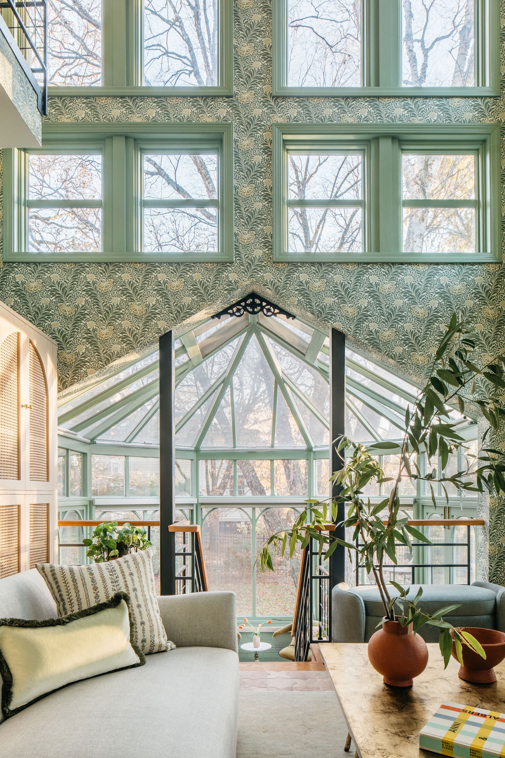 Interior view of a living room with a glass ceiling and large windows, featuring patterned wallpaper, a light-colored sofa, potted plants, and a staircase leading to a sunroom.