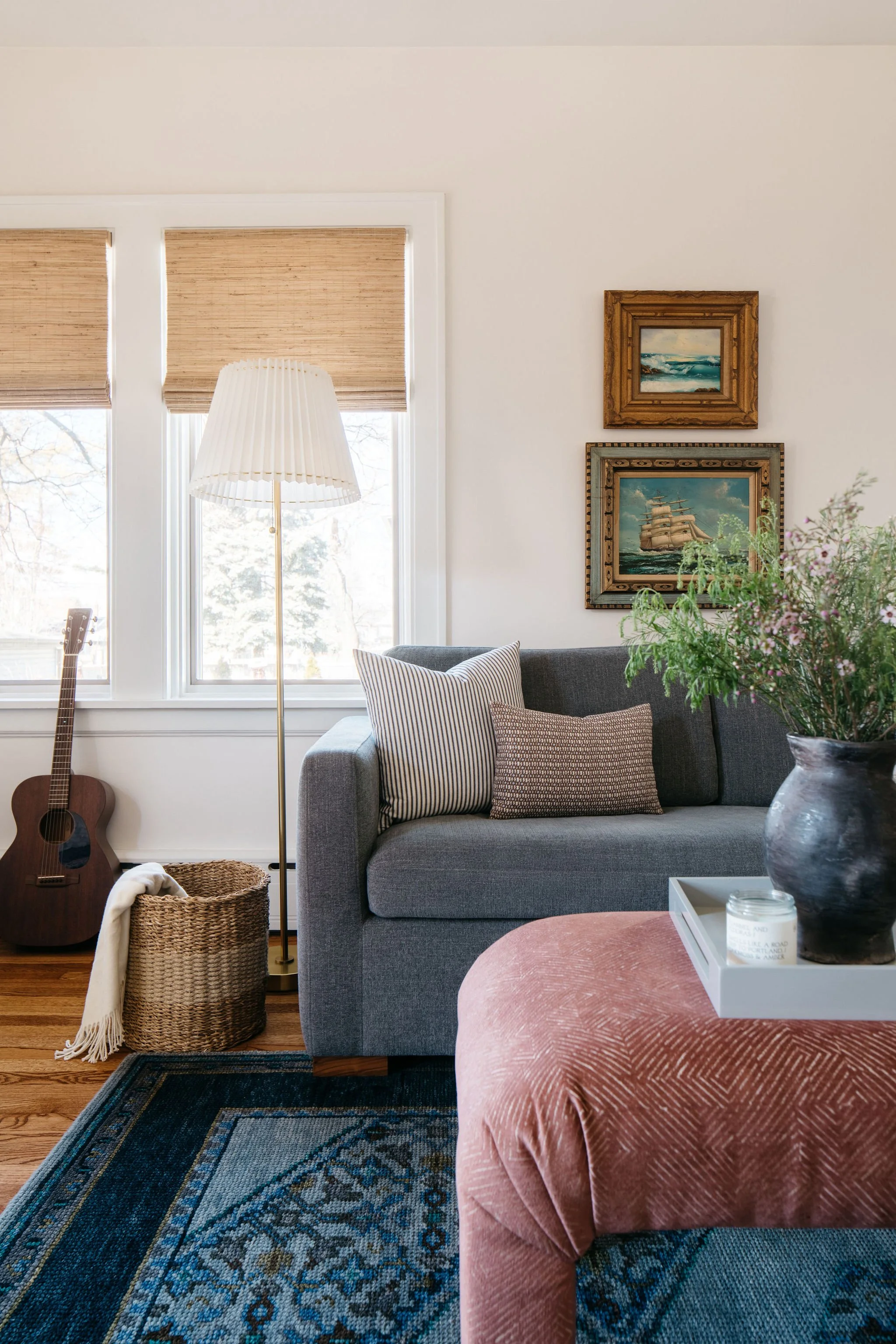 Living room with a grey sofa, decorative pillows, a tall floor lamp, framed artwork on the wall, a guitar near the window, a woven basket, and a pink upholstered ottoman with a flower vase and tray on top.