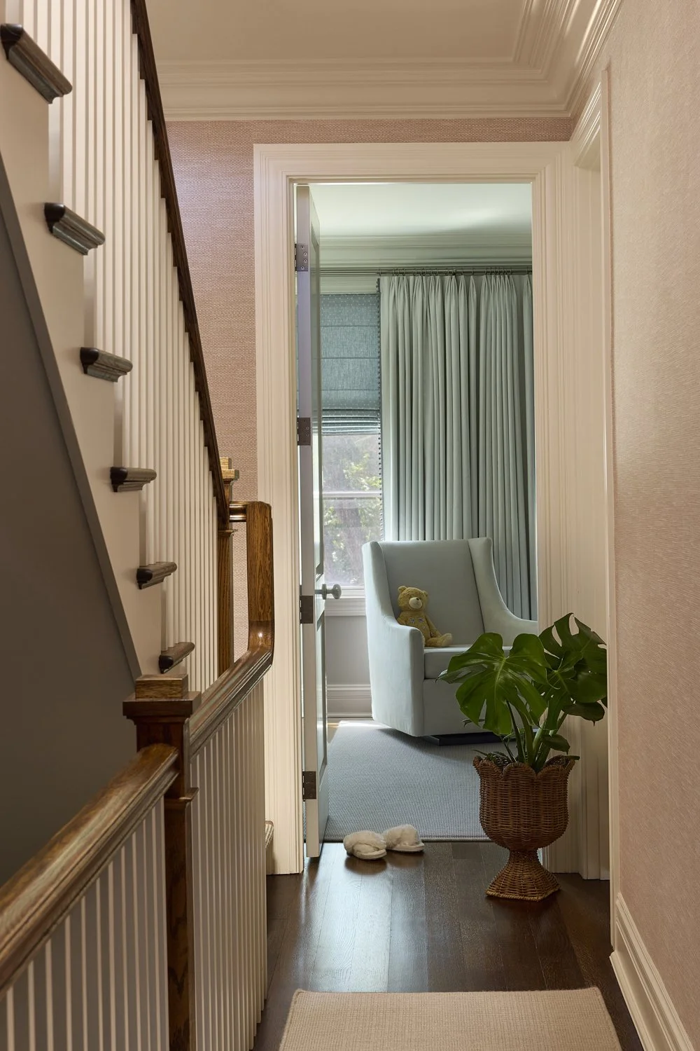 A hallway leading to a sitting area with a white armchair, a teddy bear, a large potted Monstera plant, and blue curtains in a sunlit room.