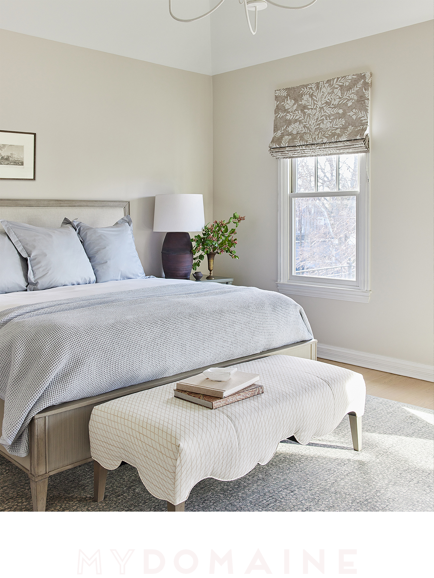 A neatly made bedroom with a bed, a window with a floral Roman shade, a bedside table with a lamp and a plant, and an upholstered bench at the foot of the bed, all in neutral tones.