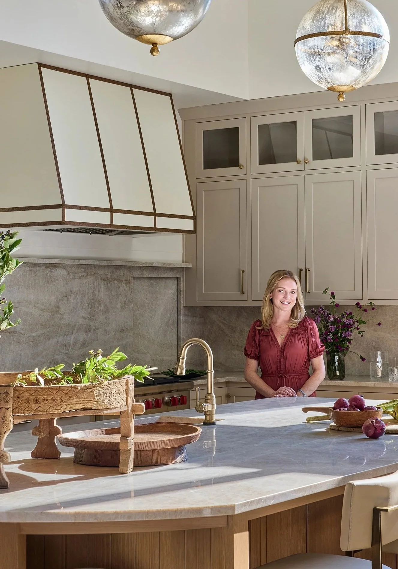 A woman with blonde hair in a red dress standing in a modern kitchen with beige cabinets, a marble island, a gold faucet, and purple flowers in a vase.