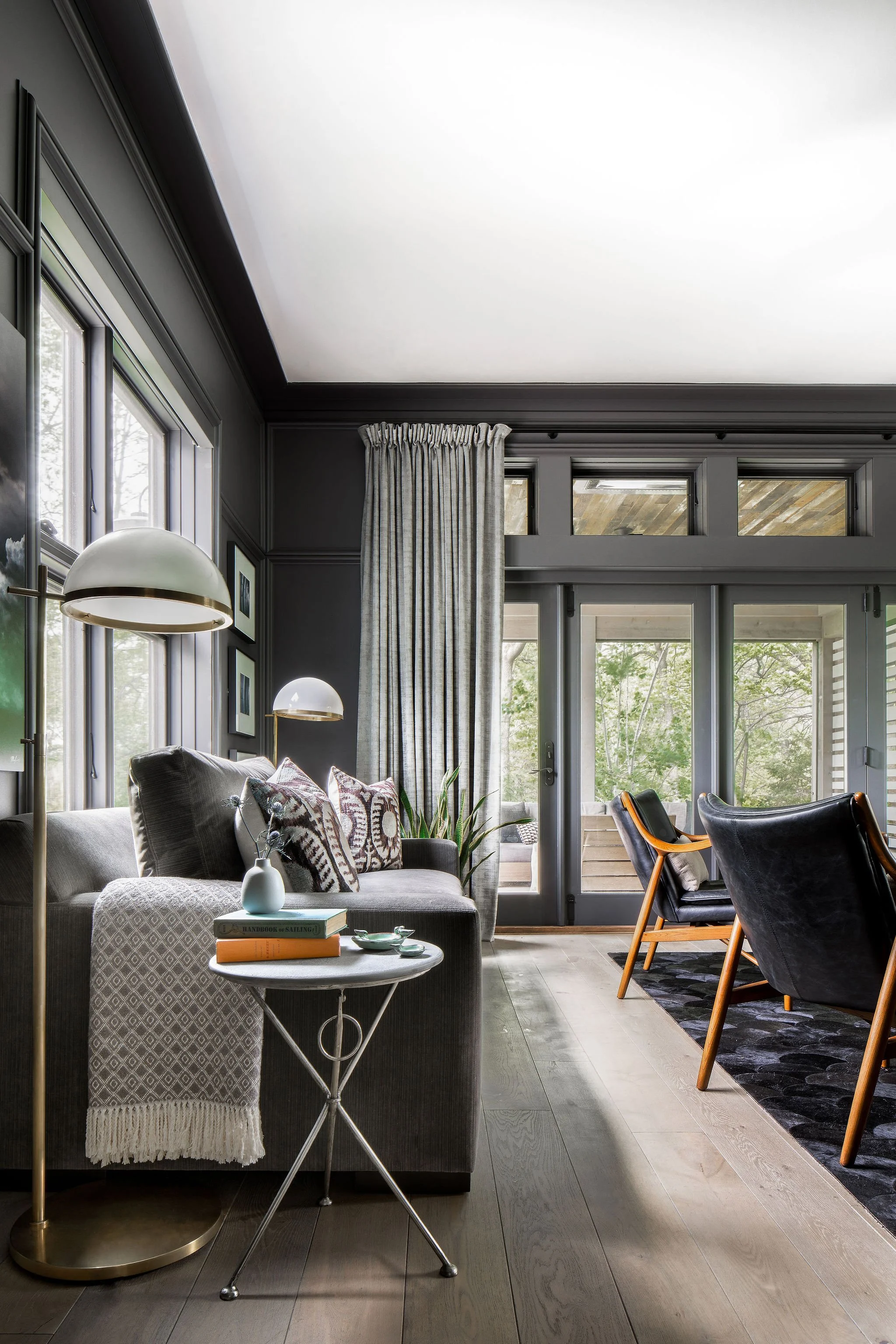 Living room with gray walls, large windows, a gray sofa decorated with patterned pillows, a small side table with books and a vase, a potted plant, and black and wooden chairs near sliding glass doors leading to an outdoor space.