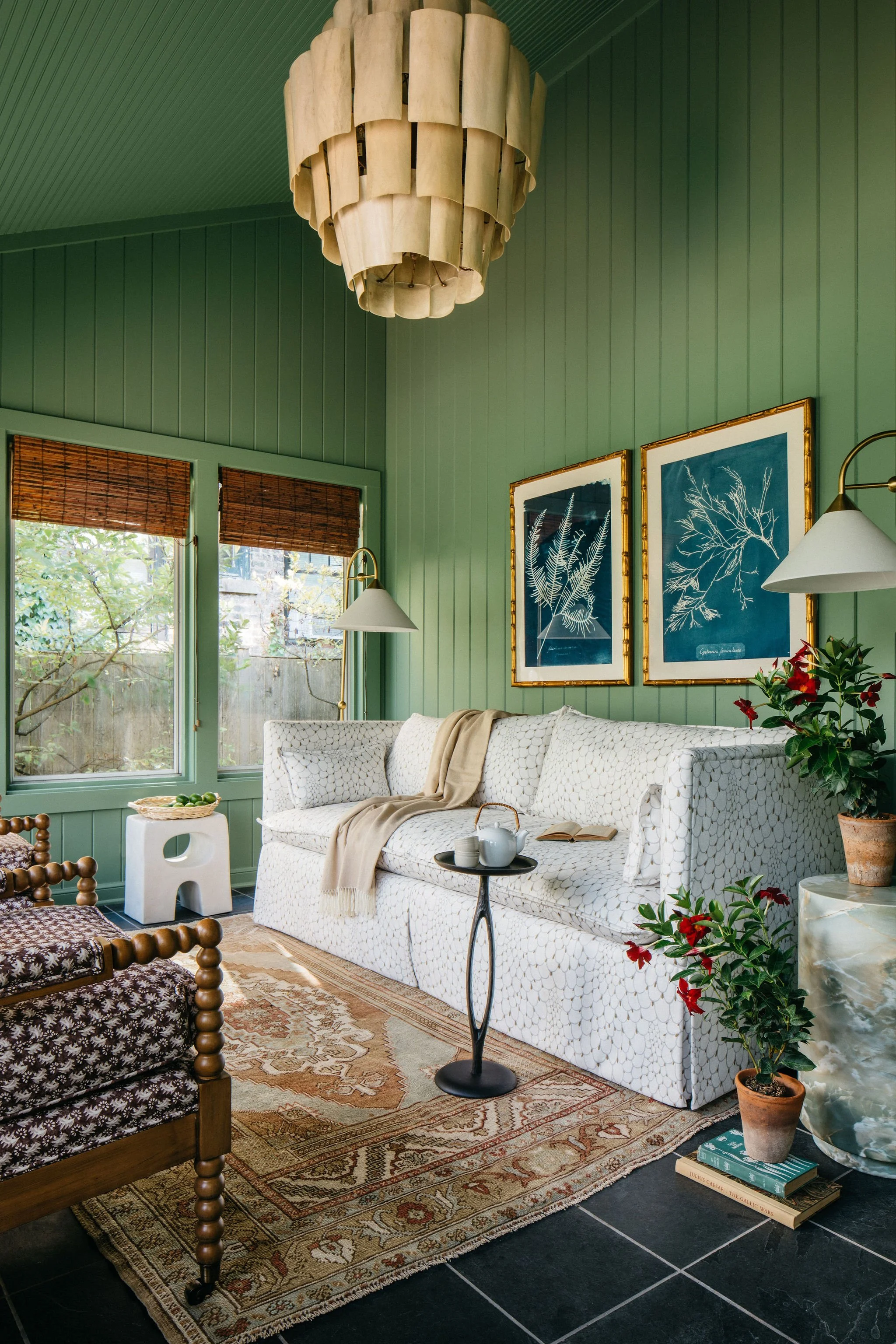 A cozy living room with green paneled walls, a white patterned sofa with a beige throw blanket, and potted plants. Two framed botanical prints hang on the wall, and there are two windows with brown blinds letting in natural light. A wooden chair with patterned fabric, a small table with teapot and cups, and a vintage rug complete the scene.