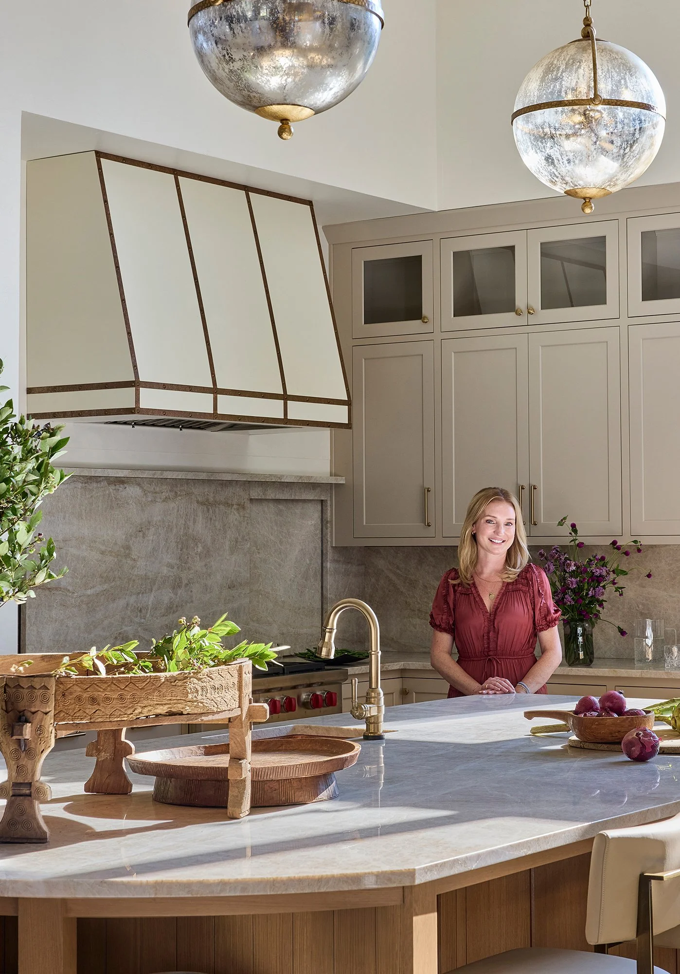 A woman smiling in a modern kitchen with beige cabinets, a marble countertop, and gold fixtures, decorated with potted plants and flowers.