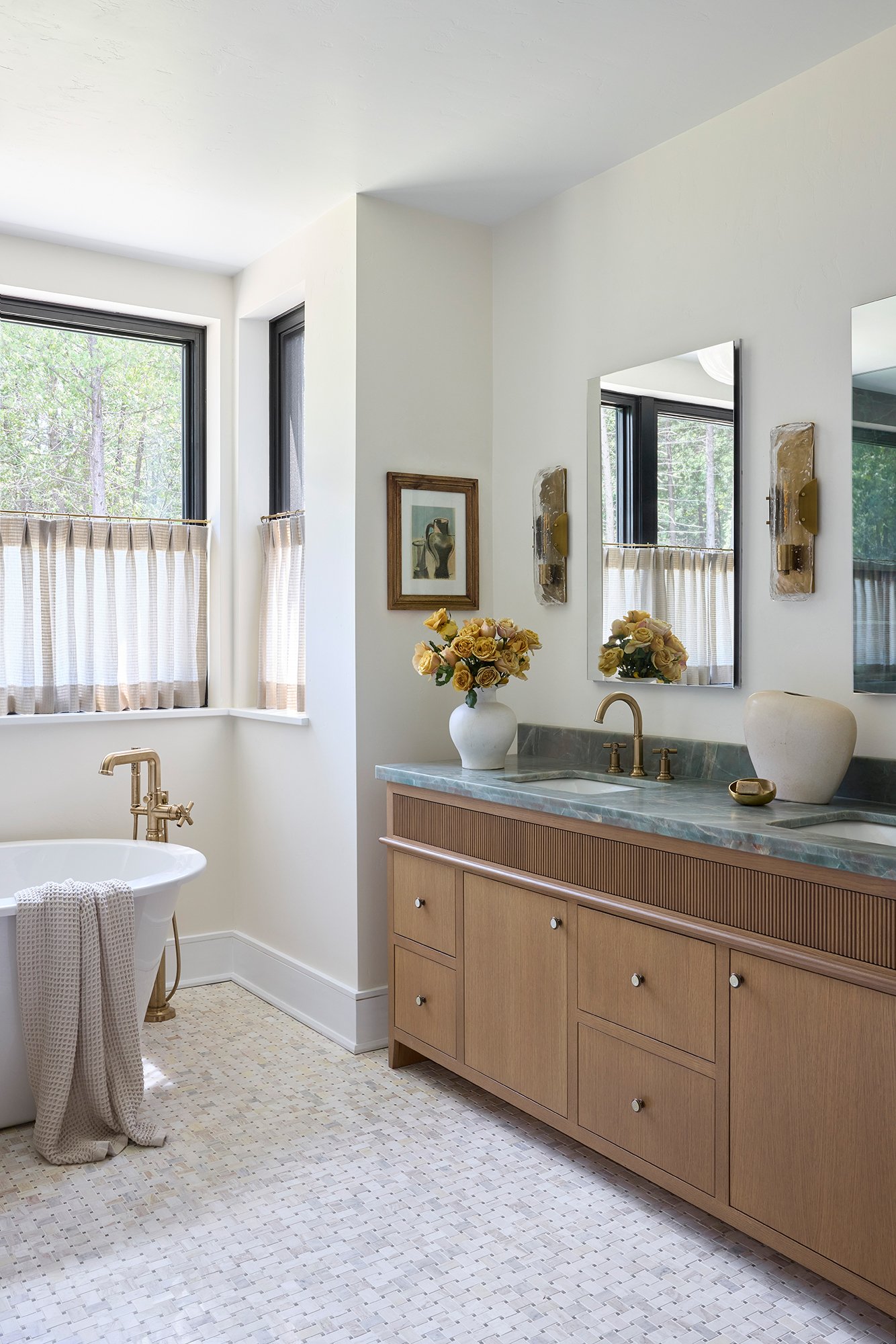 A bathroom featuring a freestanding bathtub with a gold faucet, a wooden vanity with a marble countertop, a large mirror, a vase with yellow roses, and windows with beige and white curtains.