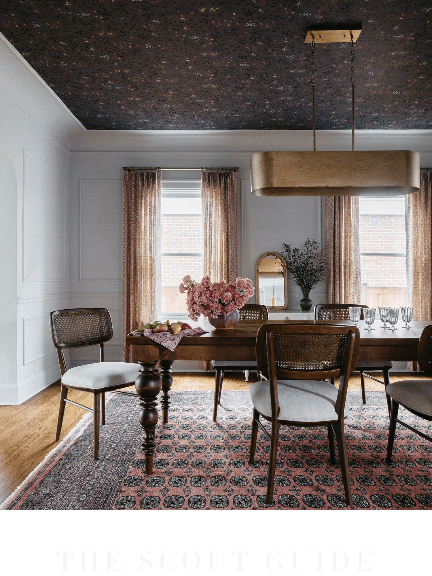 A dining room with a wooden table, six chairs, a pink floral centerpiece, and pink patterned curtains. The ceiling is dark with a colorful pattern, and a large wooden hanging light is above the table. The room has white walls, two large windows, and a decorative rug on the floor.
