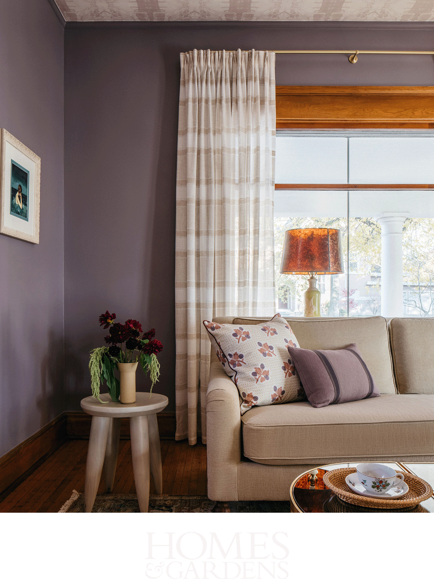 Living room with purple walls, beige sofa with floral and purple pillows, beige plaid curtains, a side table with flowers, and a window with sunlight.