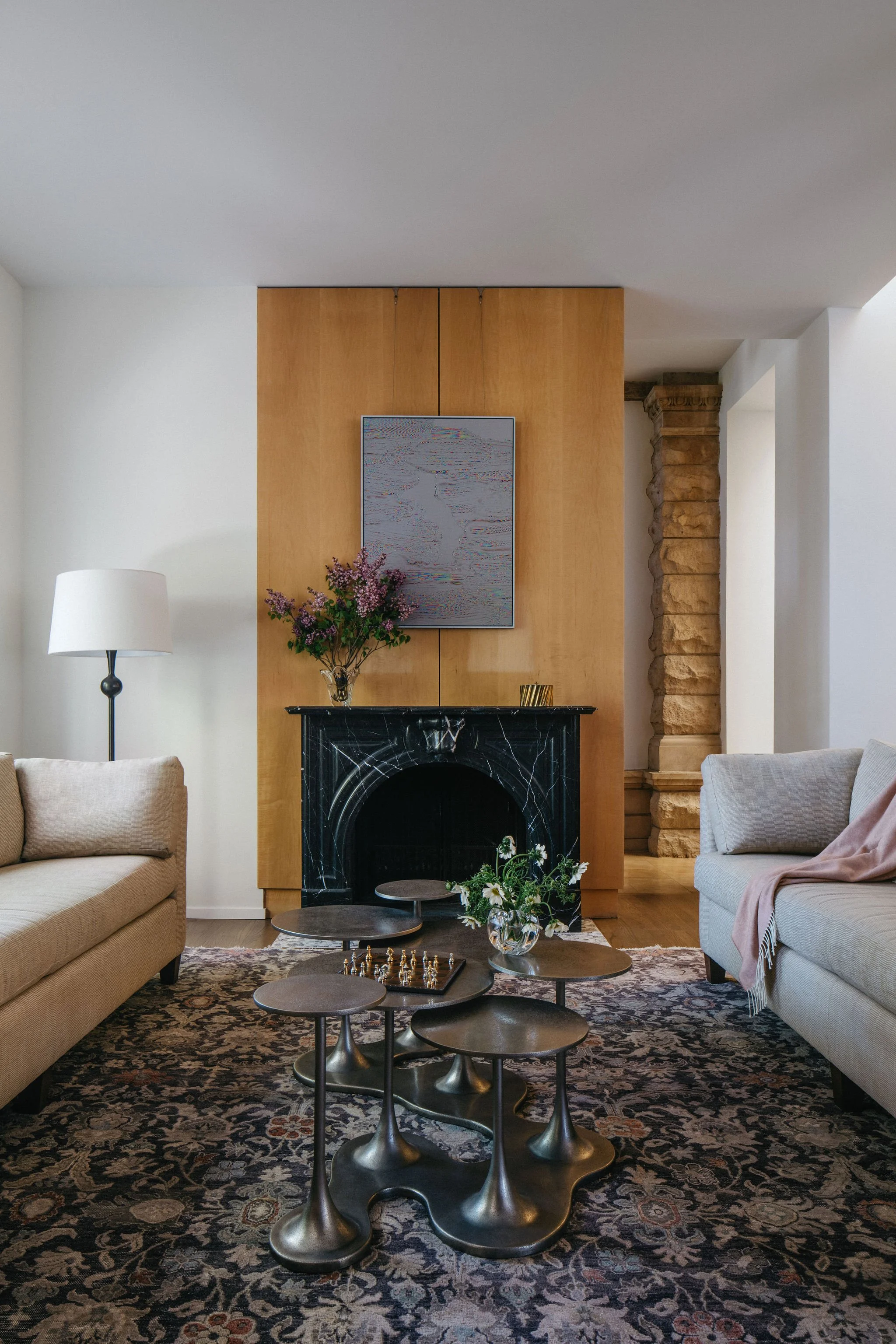 A cozy living room with beige sofas, a black marble fireplace, a vase of flowers, a modern metal coffee table, and a patterned area rug, with a floor lamp and artwork on the wall.