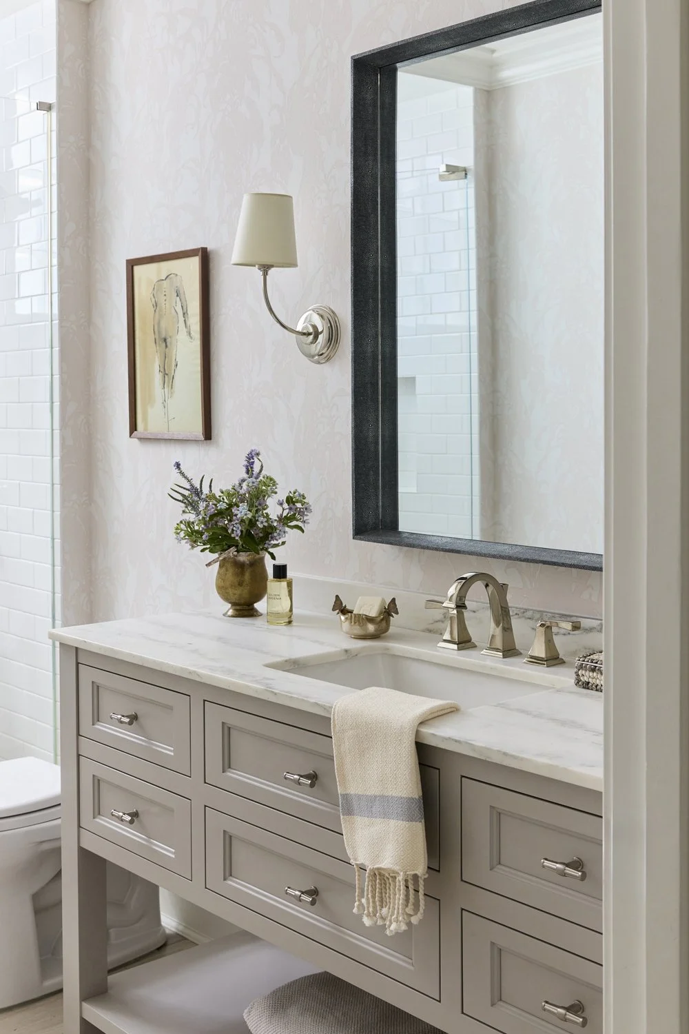 Bathroom vanity with a beige cabinet, marble countertop, metallic fixtures, a large rectangular mirror, a wall sconce, a picture of a horse, a vase with flowers, a bottle of perfume, and a folded towel. Part of a shower with white tiles is visible.