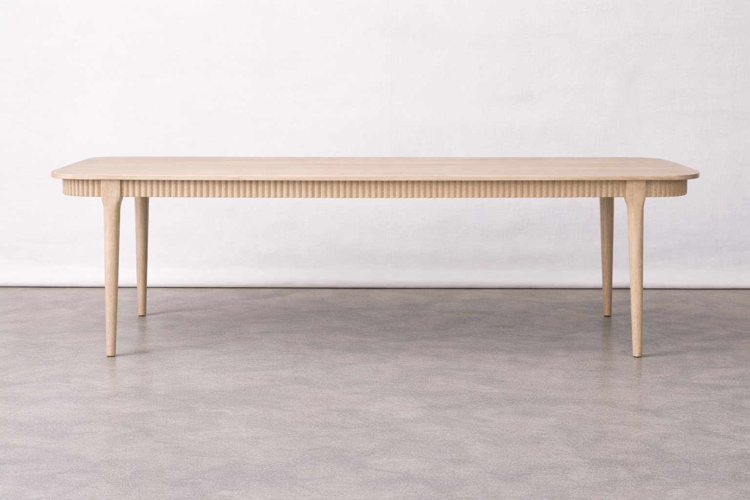 A minimalist wooden table with tapered legs and a textured, ribbed edge sitting on a concrete floor against a plain white wall.
