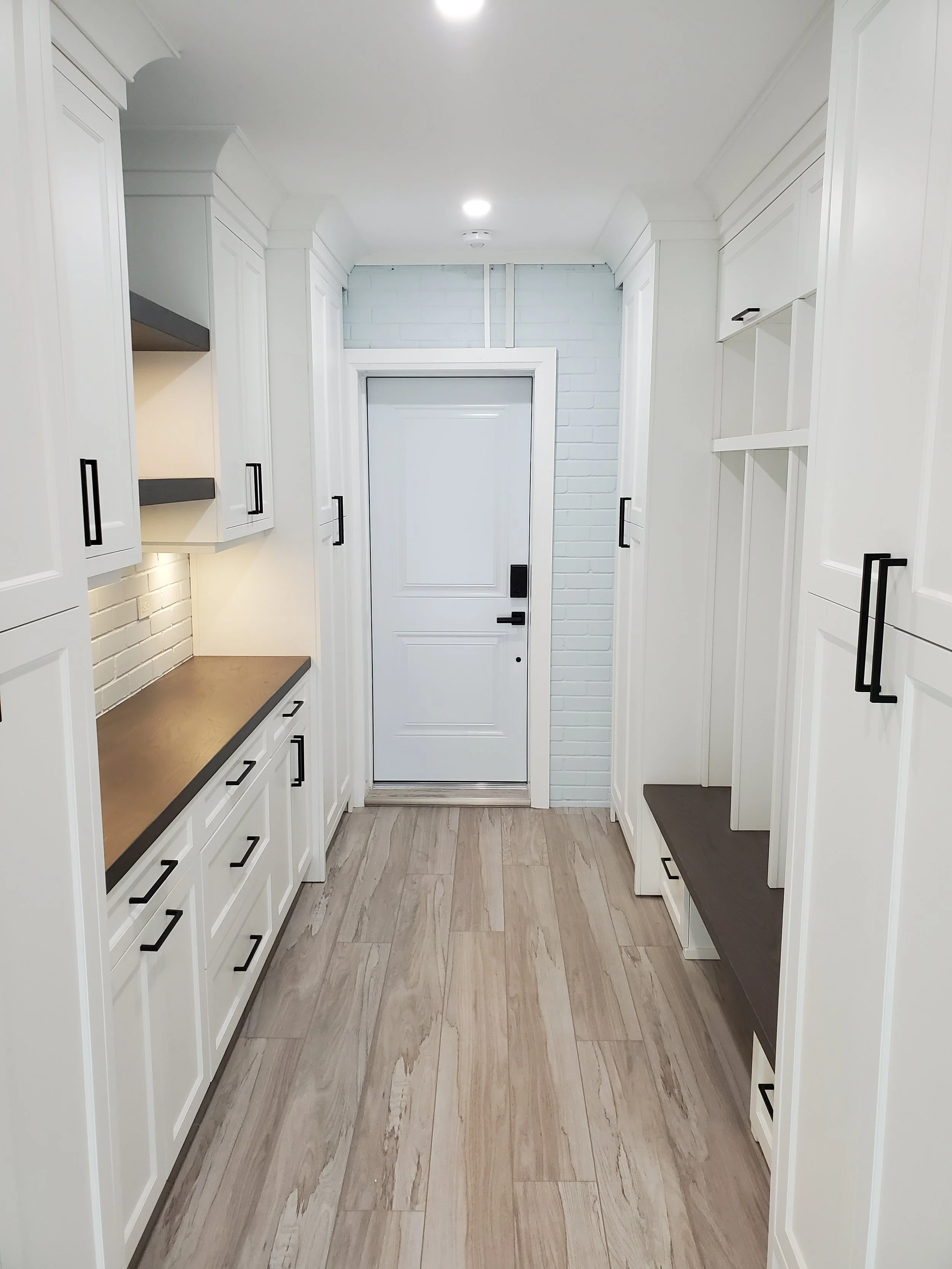 Modern laundry or mudroom with white cabinets, black handles, wood-look flooring, and a small countertop on the left side. White brick wall behind the door, with additional storage on the right side.