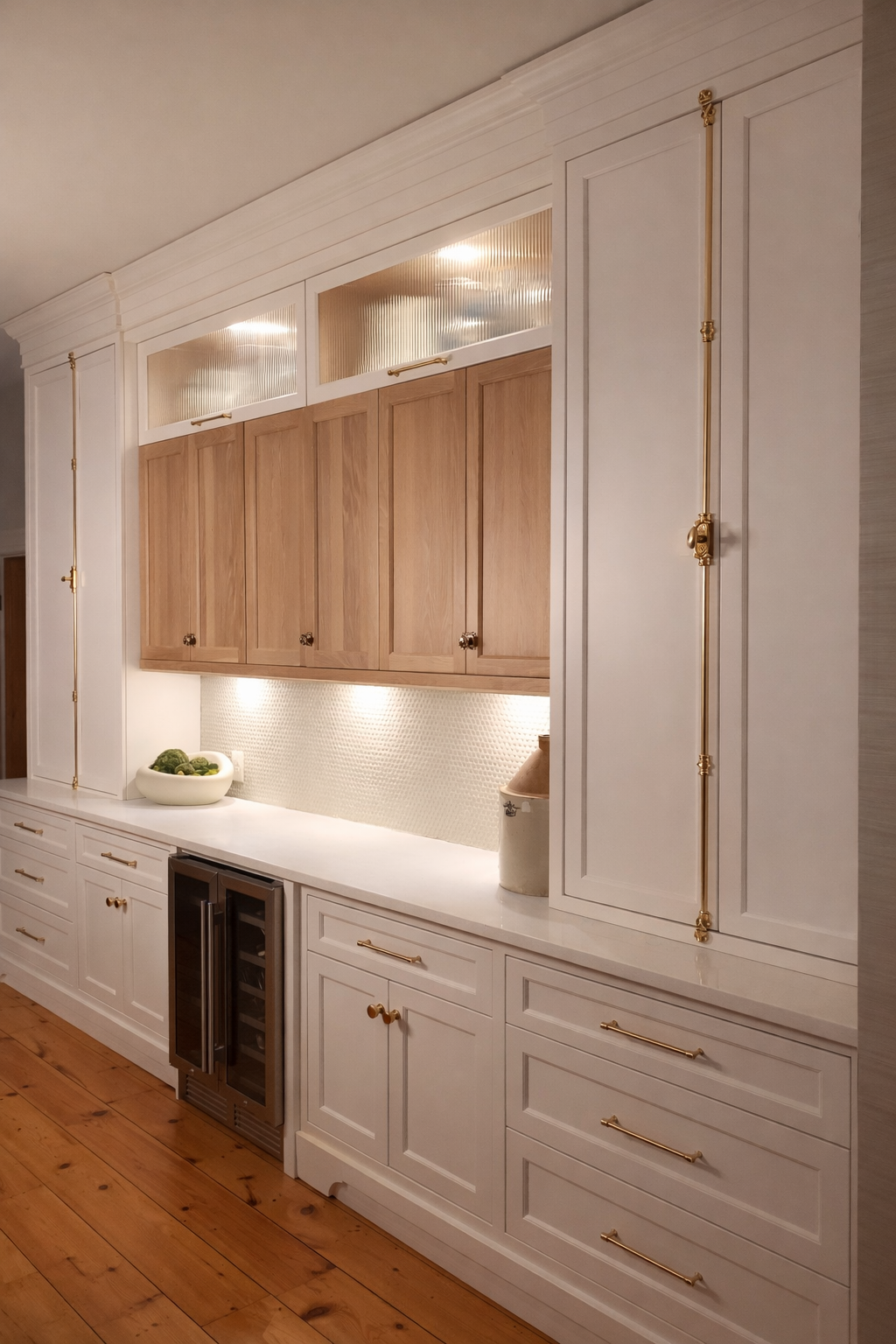 A kitchen with white and wooden cabinets, gold handles, and a wood floor. There is a built-in wine cooler and a bowl of green vegetables on the countertop.