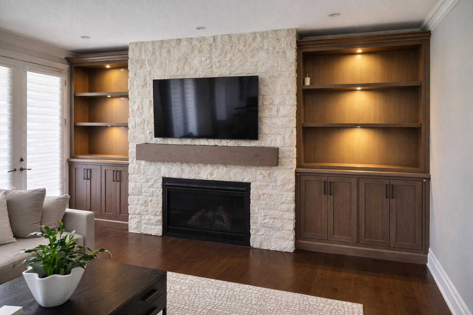 Living room featuring a stone fireplace with a mounted flat-screen TV, wooden built-in shelves with display lighting, and a beige sofa with a potted plant on a dark coffee table.
