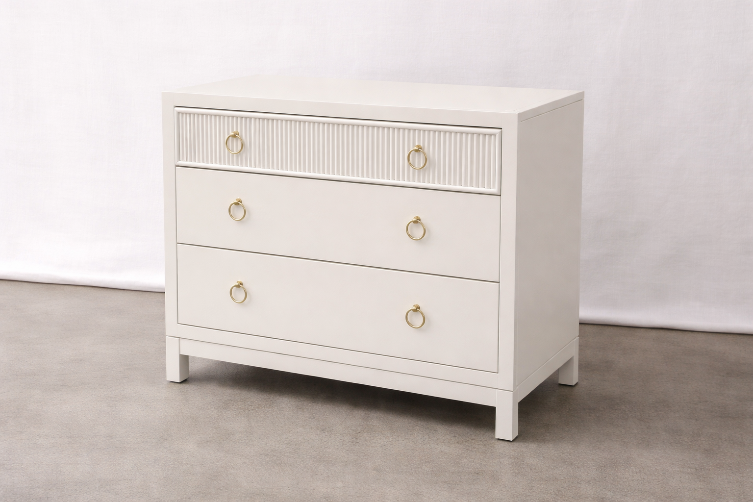 White dresser with four drawers, gold ring handles, and a textured front panel on the top drawer, placed on a gray carpeted floor against a light background.