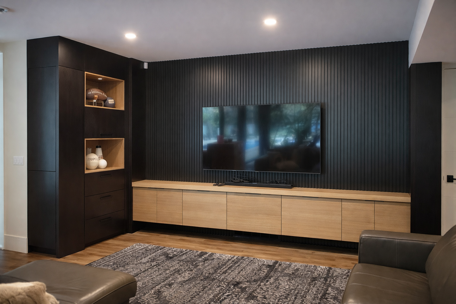 Modern living room with a large flat-screen TV mounted on a black textured accent wall, black and wood shelving and cabinetry, a Persian rug, and leather furniture.