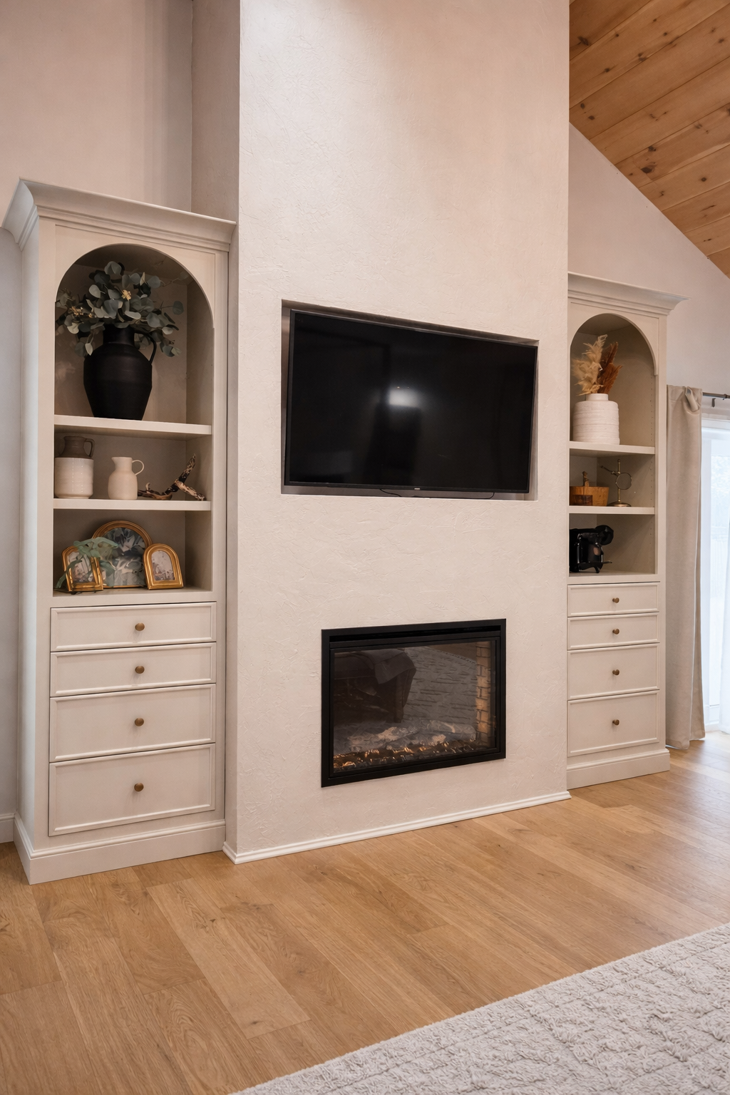 Living room with a fireplace below a wall-mounted TV, flanked by two white built-in cabinets with decorative items and drawers, on a wooden floor with a rug partially visible.