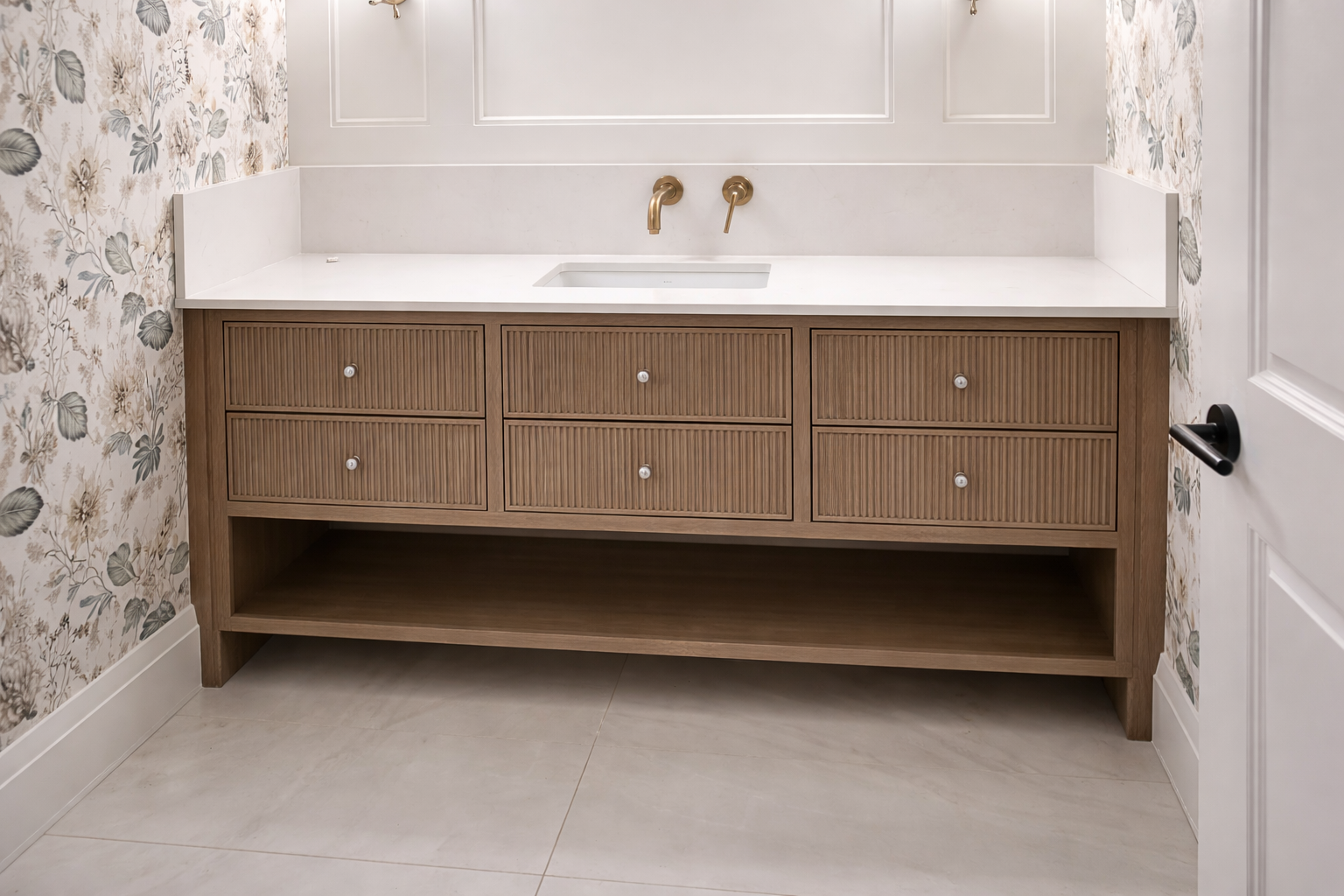 Bathroom vanity with a wooden cabinet, white countertop, small sink, and gold faucet, against floral wallpaper and white wall paneling.
