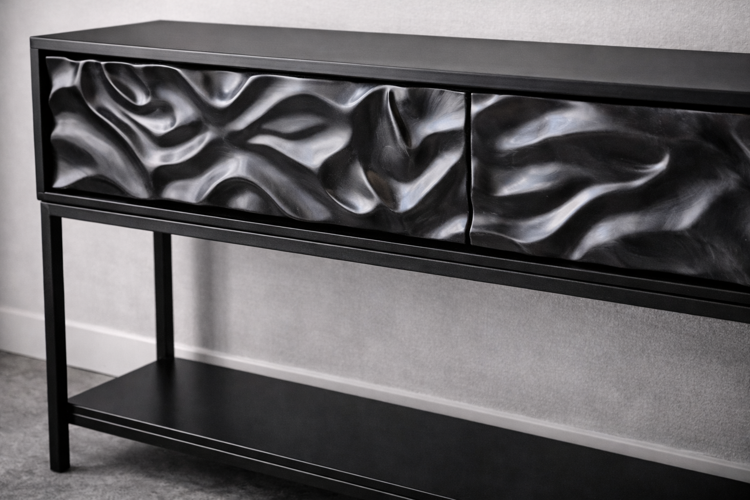 A modern black console table with a textured, wavy, metallic-designed drawer front and a lower shelf, placed against a lightly textured wall.