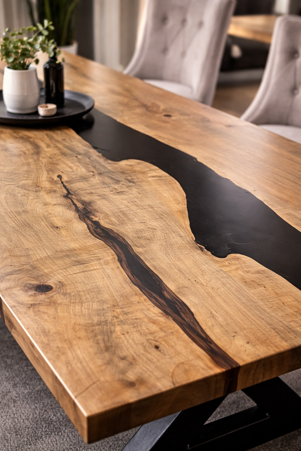 Close-up of a wooden dining table with a natural finish and a black epoxy resin river running through the center, surrounded by beige upholstered chairs with tufted backs.
