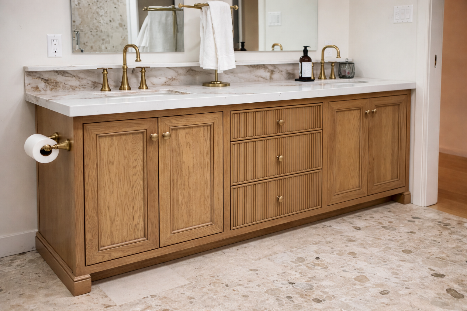 Bathroom vanity with wooden cabinets, marble countertop, two gold faucets, soap dispenser, mirror, and a towel. Toilet paper holder on the side, soap and glass on the right, wall outlets behind the sink.