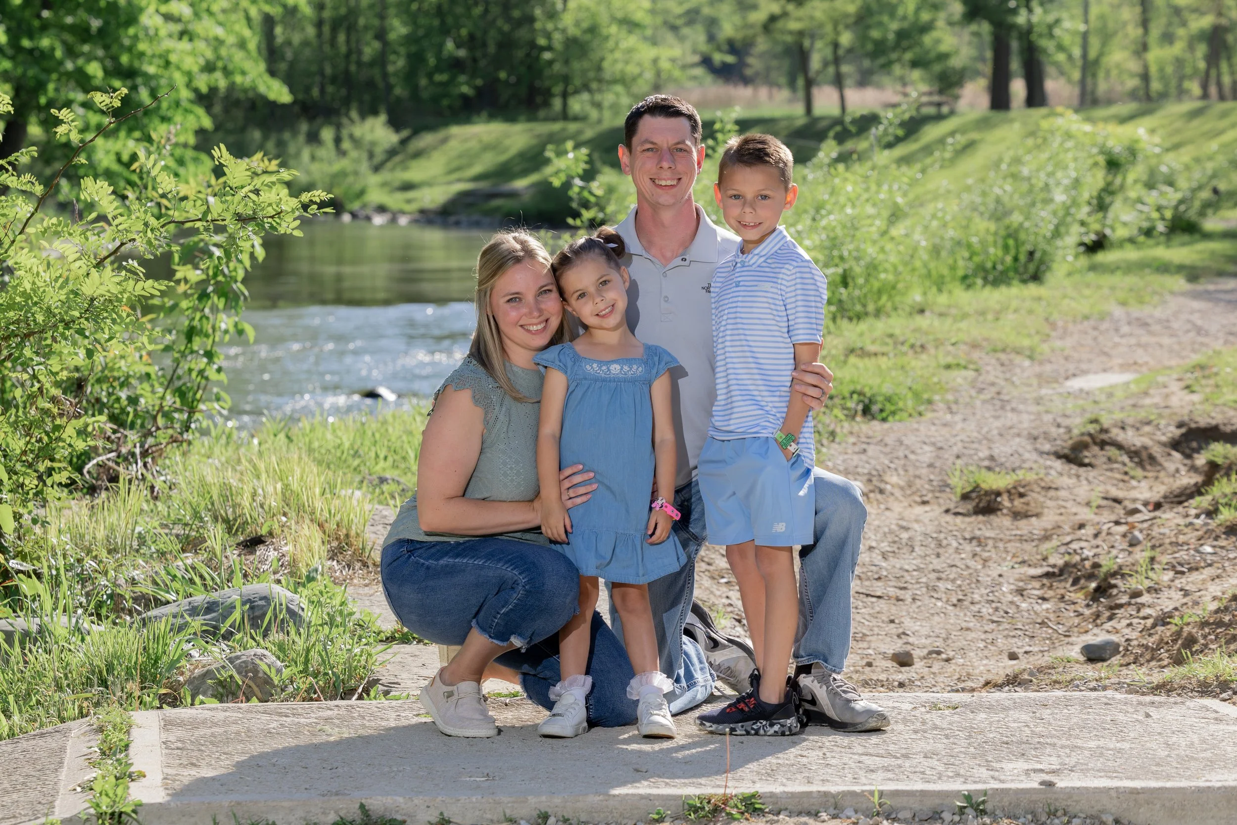 A family of four standing outdoors near a creek with lush trees and greenery in the background, smiling at the camera.