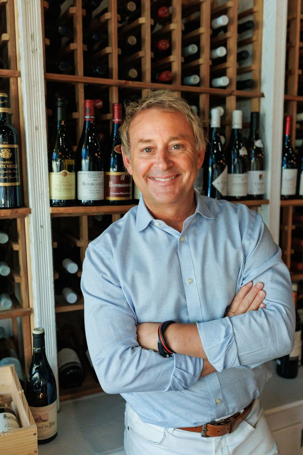 Smiling man in a light blue shirt with arms crossed, standing in front of a wine rack with bottles.