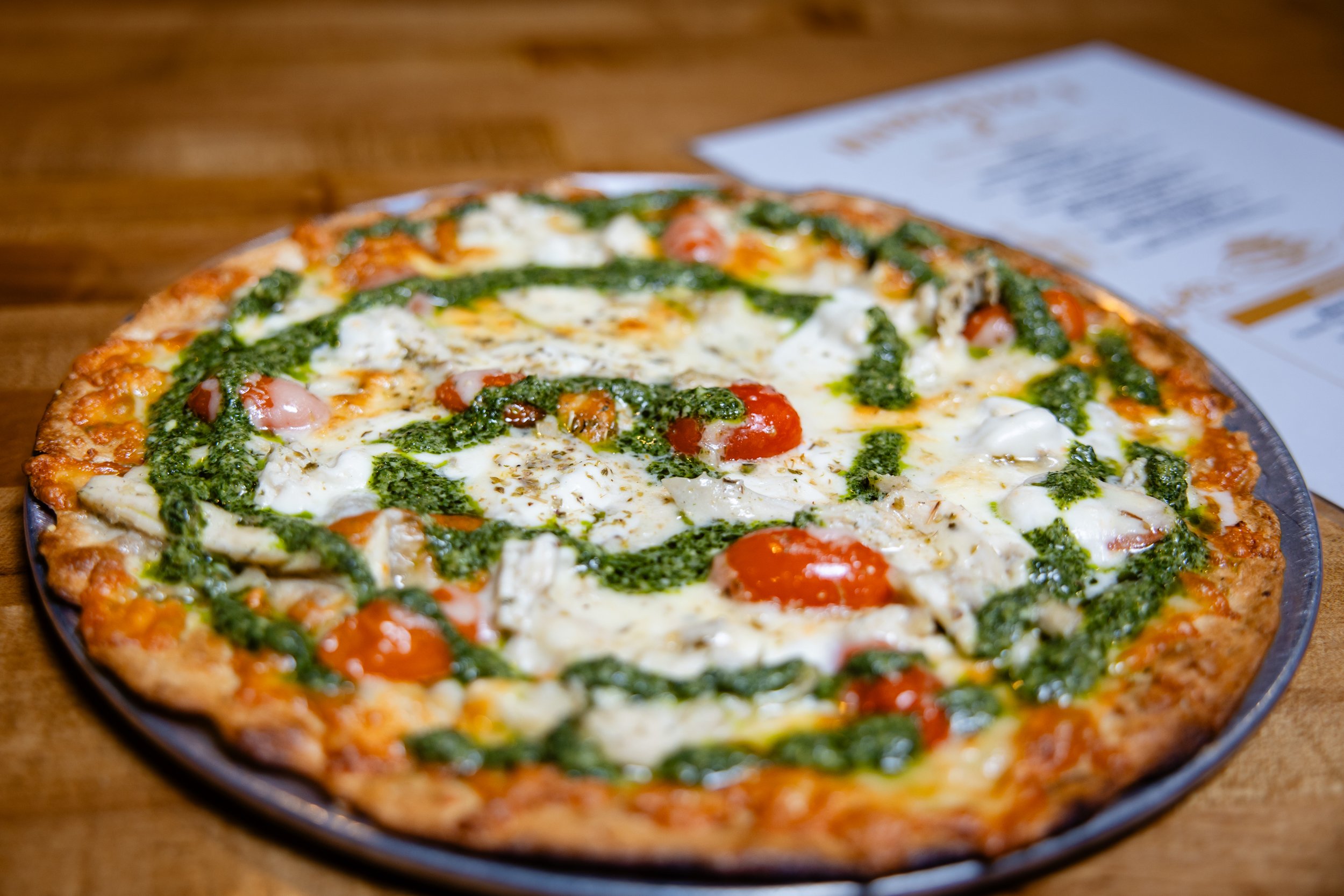 Close-up of a pizza with cherry tomatoes, basil pesto, and white cheese on a wooden table with a receipt nearby.