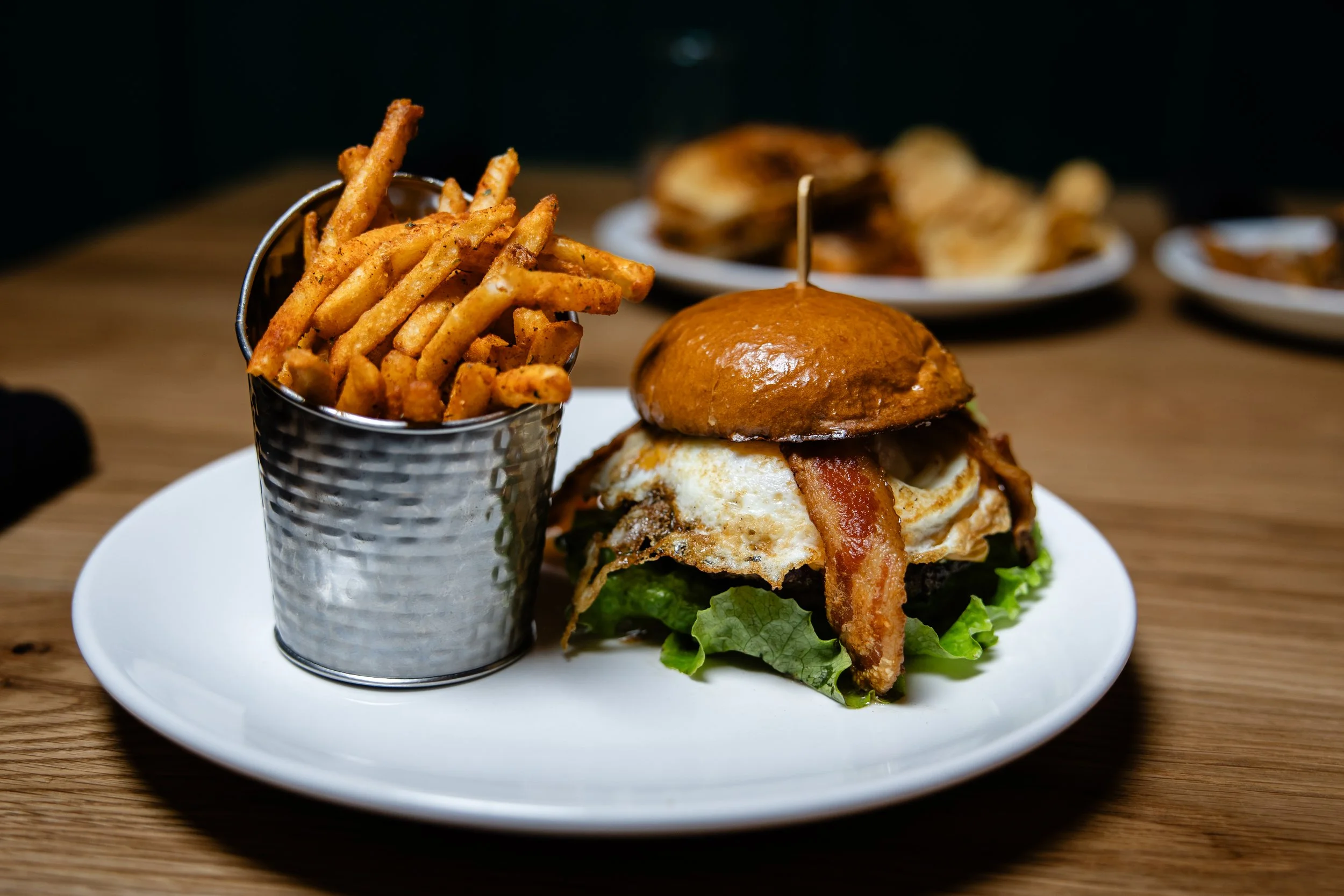 A plate with a burger topped with bacon, fried egg, and lettuce, accompanied by a side of French fries in a metal cup on a wooden table.