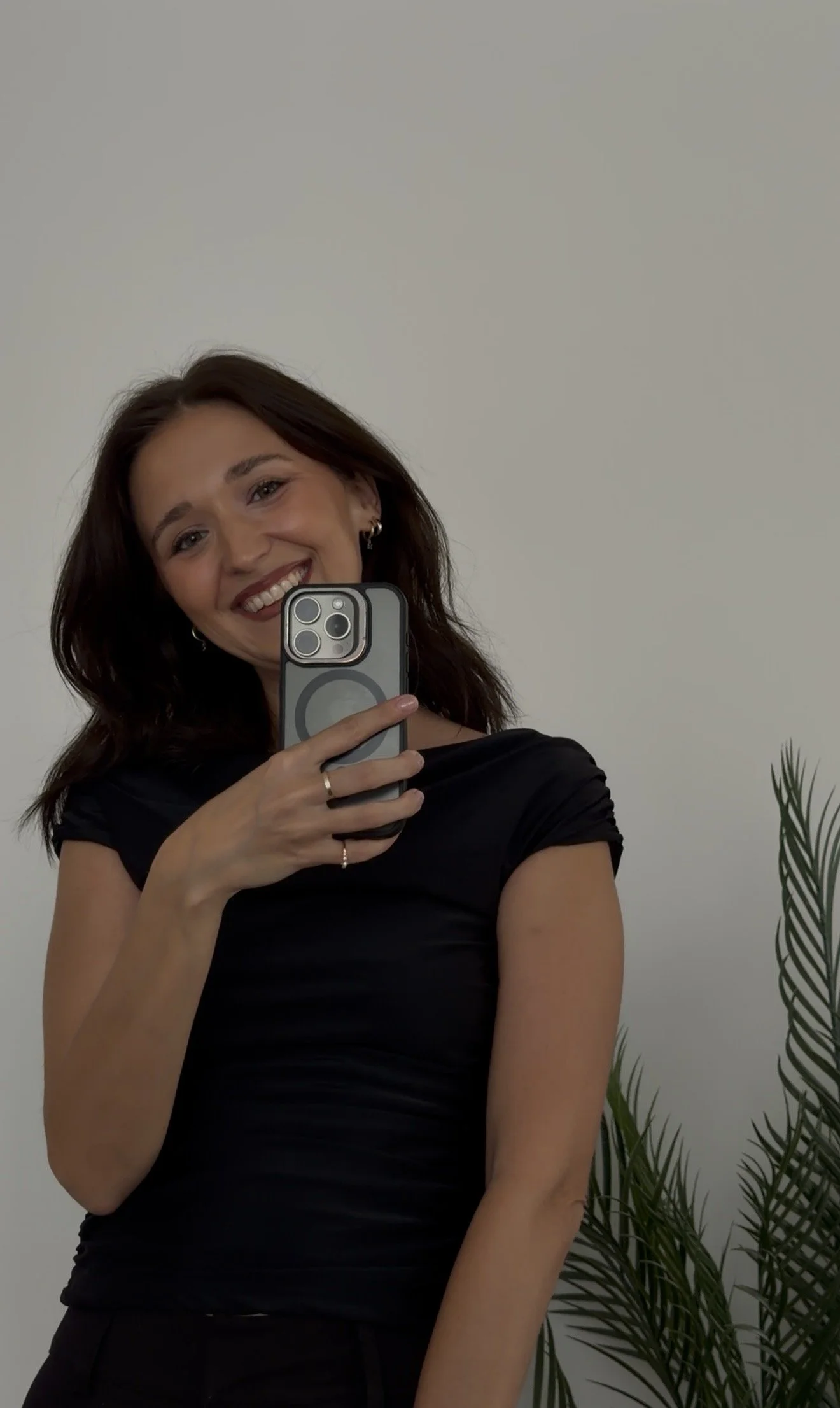 A woman with shoulder-length dark brown hair smiling and taking a selfie with a smartphone. She is wearing a black top and has a plant with long, narrow leaves in the background.