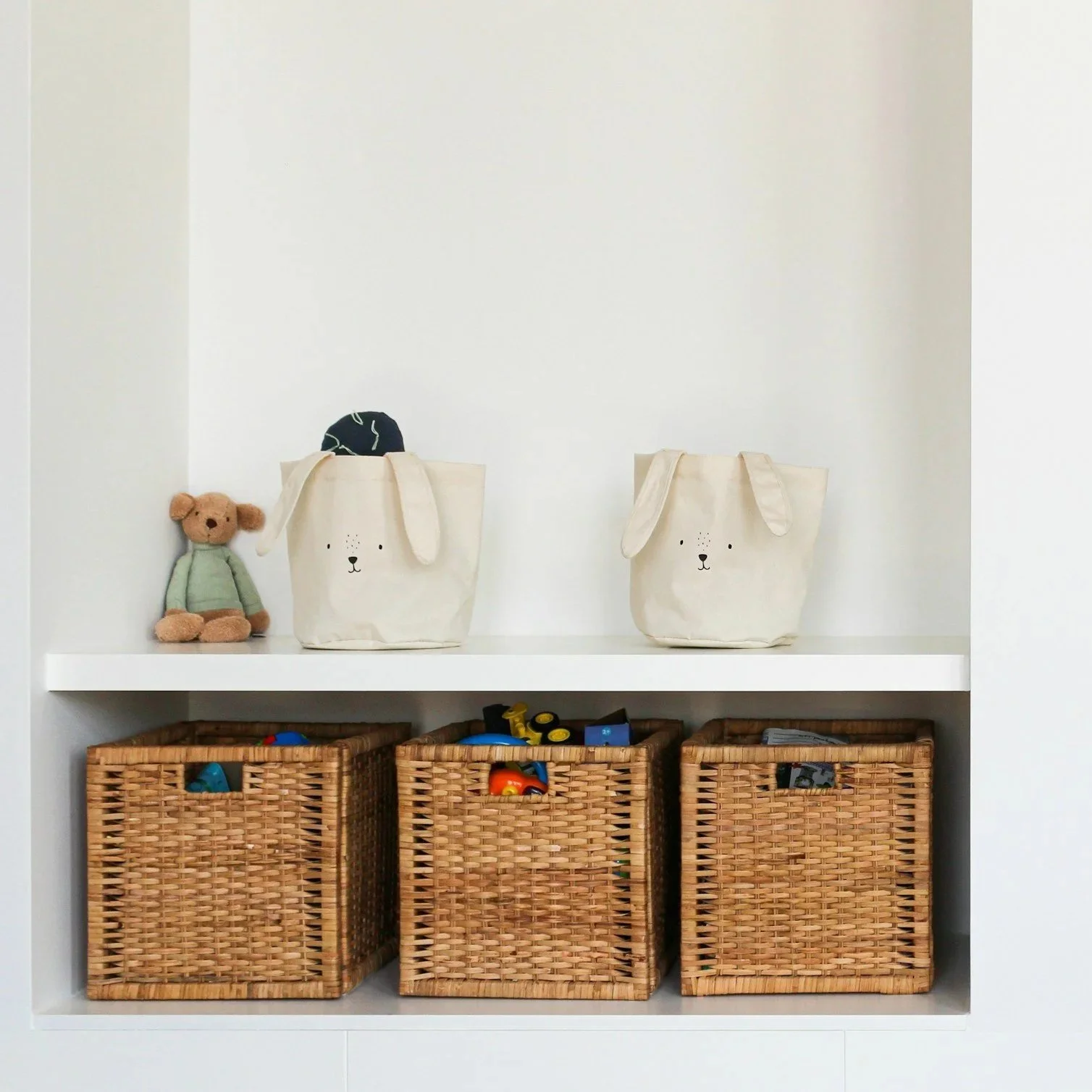 A white shelf with two woven baskets on the bottom shelf filled with toys, two white tote bags with dog face designs on the top shelf, and a small stuffed bear with a green shirt on the left side of the top shelf.