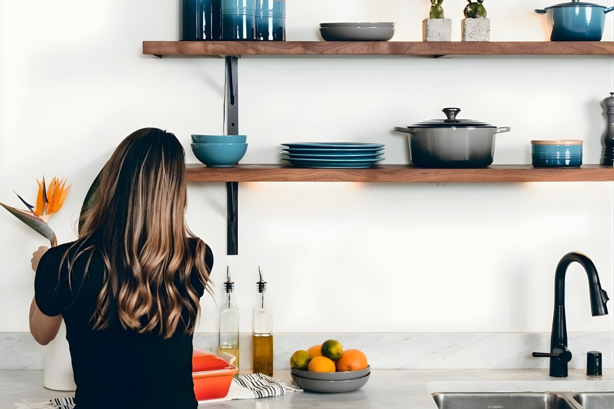 A woman with long wavy hair standing in a kitchen, holding a bird of paradise flower. The kitchen has wooden shelves with blue and gray bowls, plates, and cookware, a sink with a black faucet, and a bowl of citrus fruits on the counter.