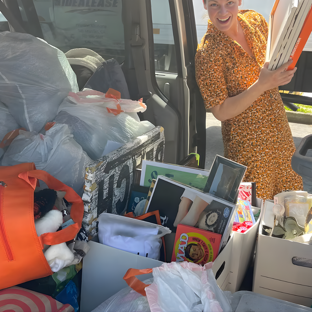 A woman in an orange patterned dress smiling and holding a clipboard, standing next to an open vehicle filled with bags, boxes, and various items for donation or sale.