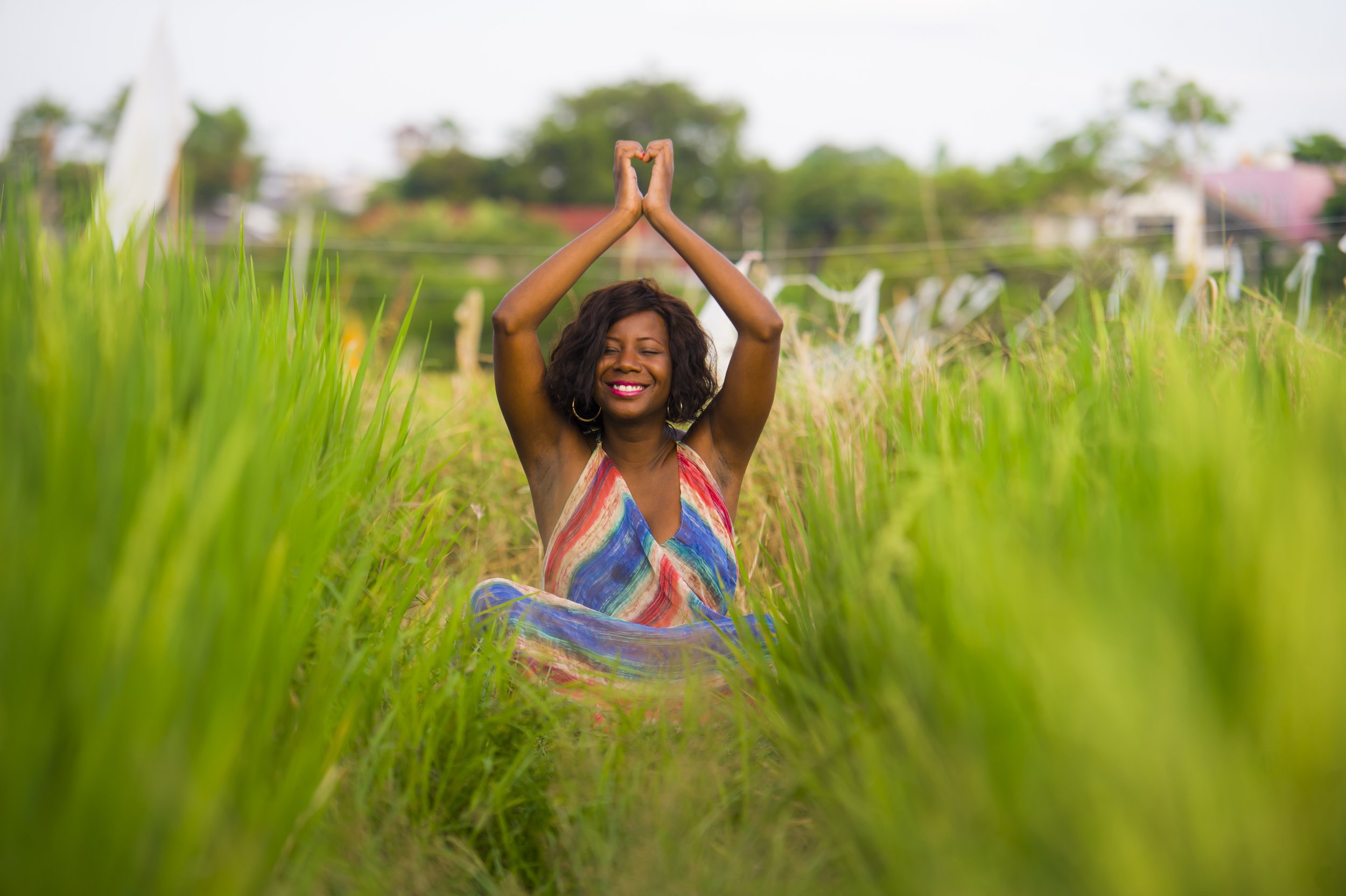 A woman with curly hair and a colorful dress sitting cross-legged in a lush green field, smiling and cultivating self-compassion and internal safety. Increasing emotional regulation and resilience.