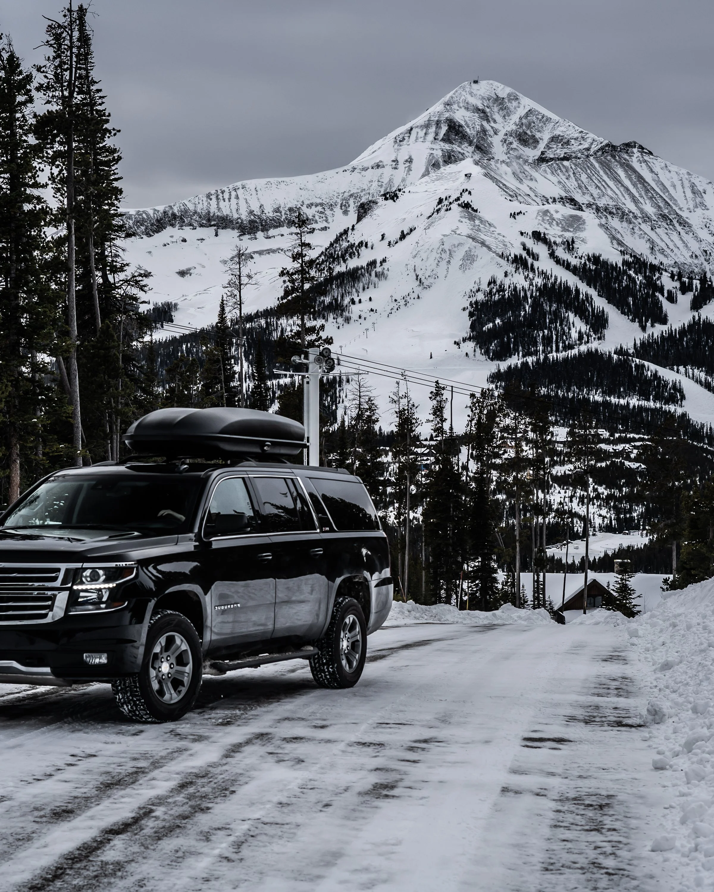 A black SUV with a rooftop cargo box parked on a snowy mountain road, with snow-covered pine trees and a large snow-capped mountain in the background.
