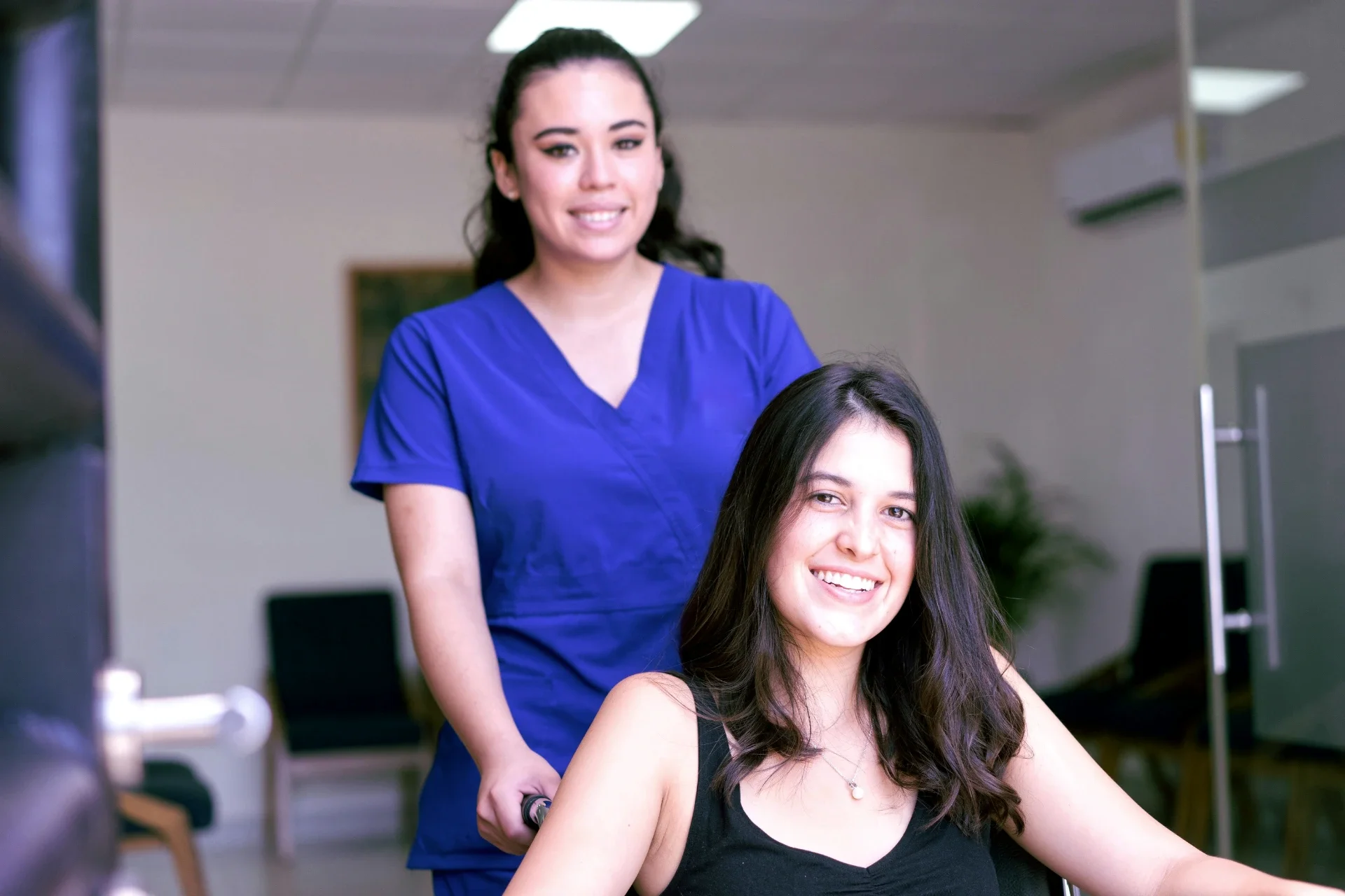 A woman in a blue medical scrub standing behind a woman with long dark hair smiling while sitting in a wheelchair.