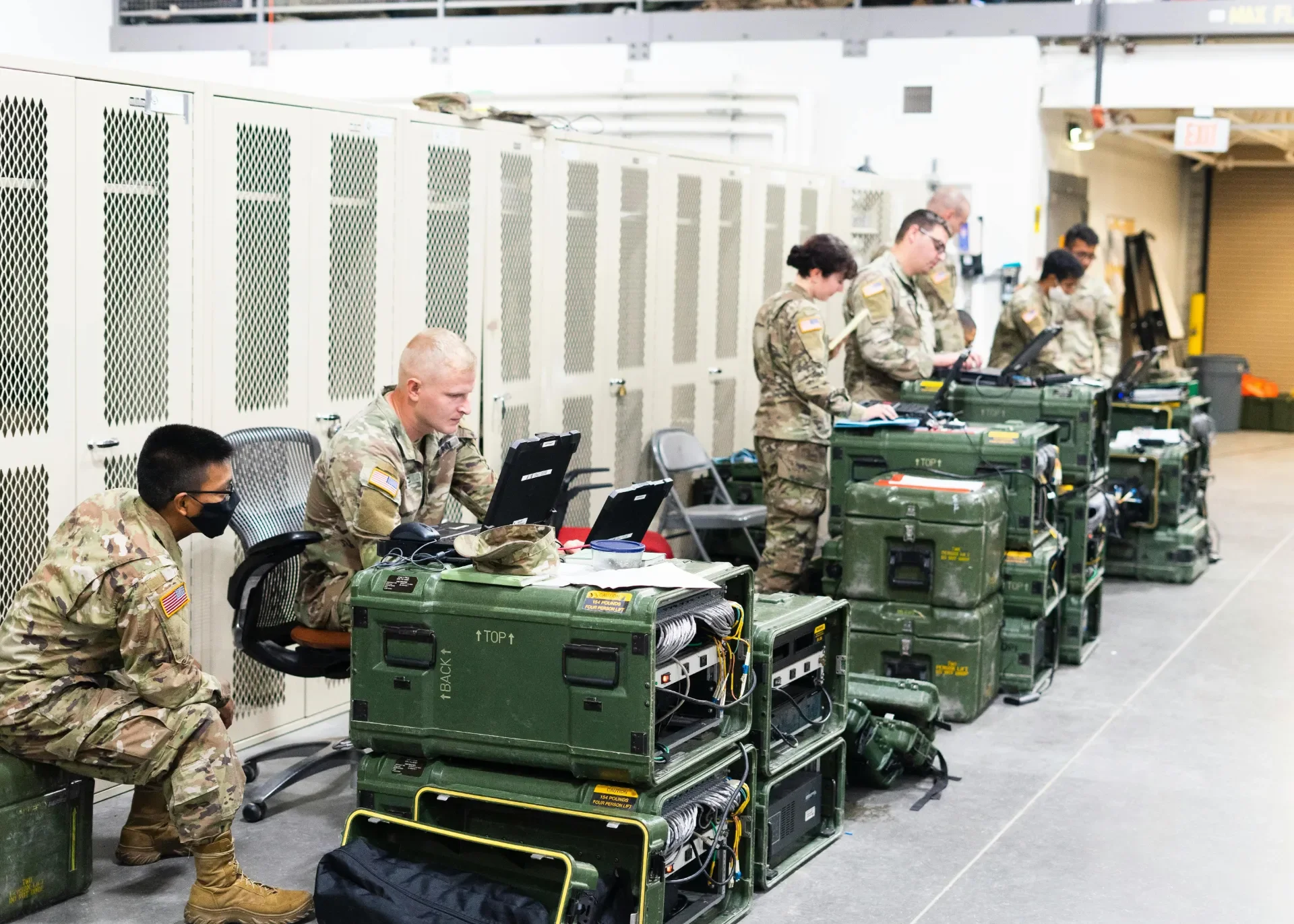 Military personnel working on laptops and communication equipment in a room with lockers.