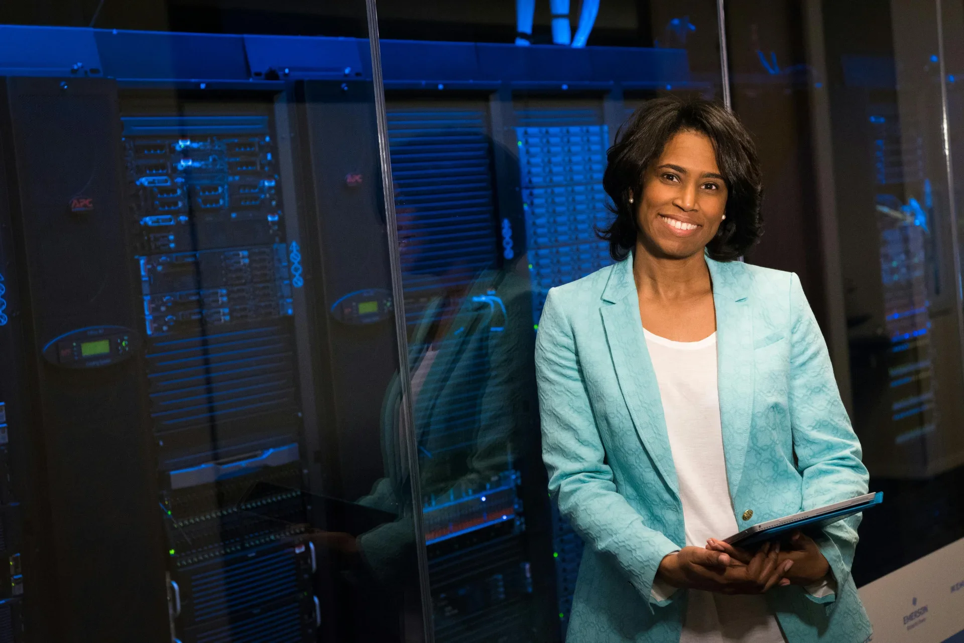 A woman in a light teal blazer and white shirt holding a tablet, standing in front of server racks in a data center or server room.