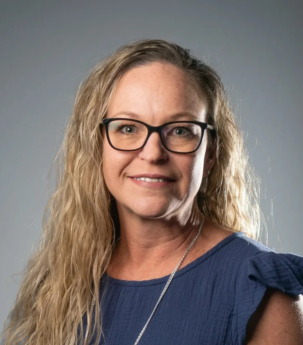A woman with blonde, wavy hair wearing glasses and a blue dress with ruffled shoulders, smiling against a neutral gray background.