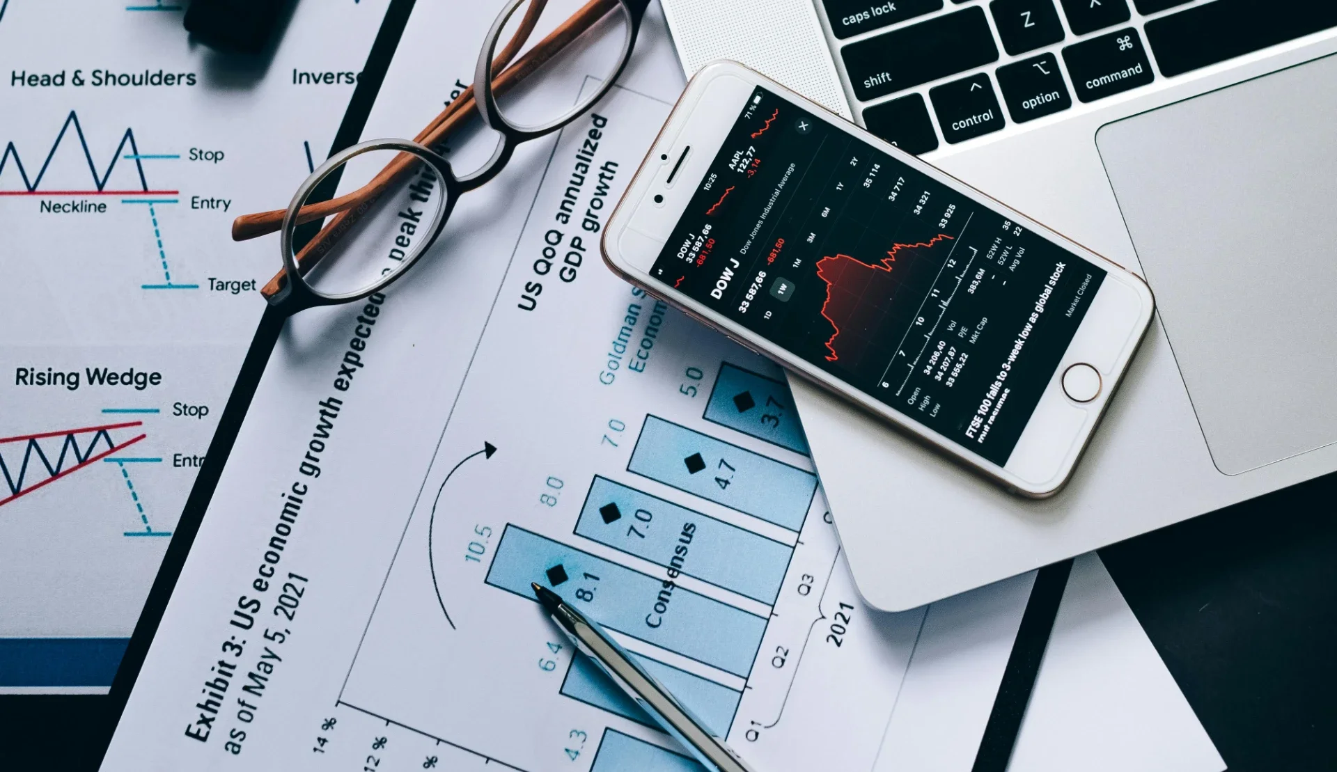 Close-up of financial charts, graphs, a smartphone displaying stock market data, a laptop, a pair of glasses, and a pen on a desk.