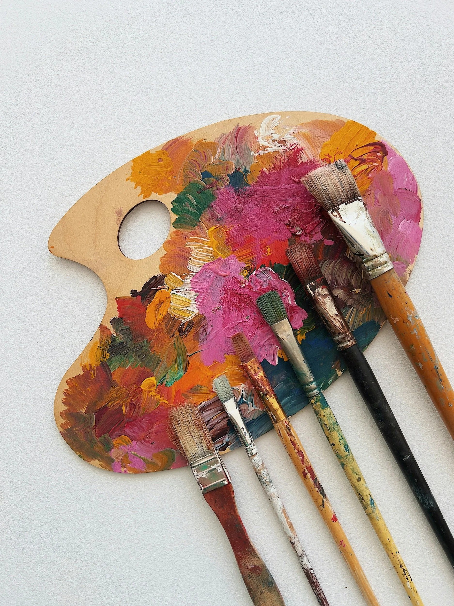 Wooden paint palette with multiple paintbrushes resting on it, all covered in colourful acrylic paint on a white background.