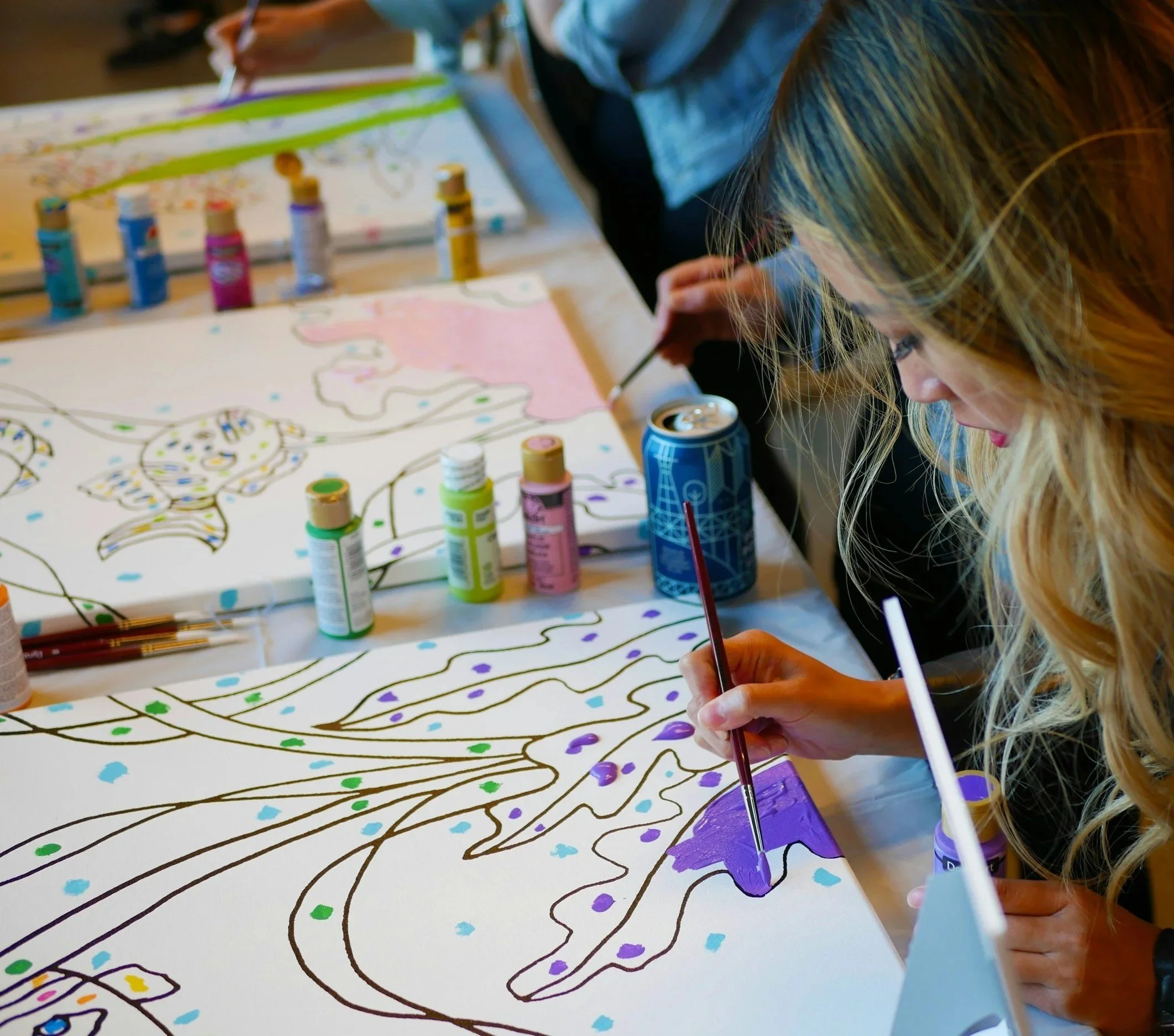 A side view of a person painting a large, colourful page with markers and paint on canvas on a table. You can see others working on the same table behind them.