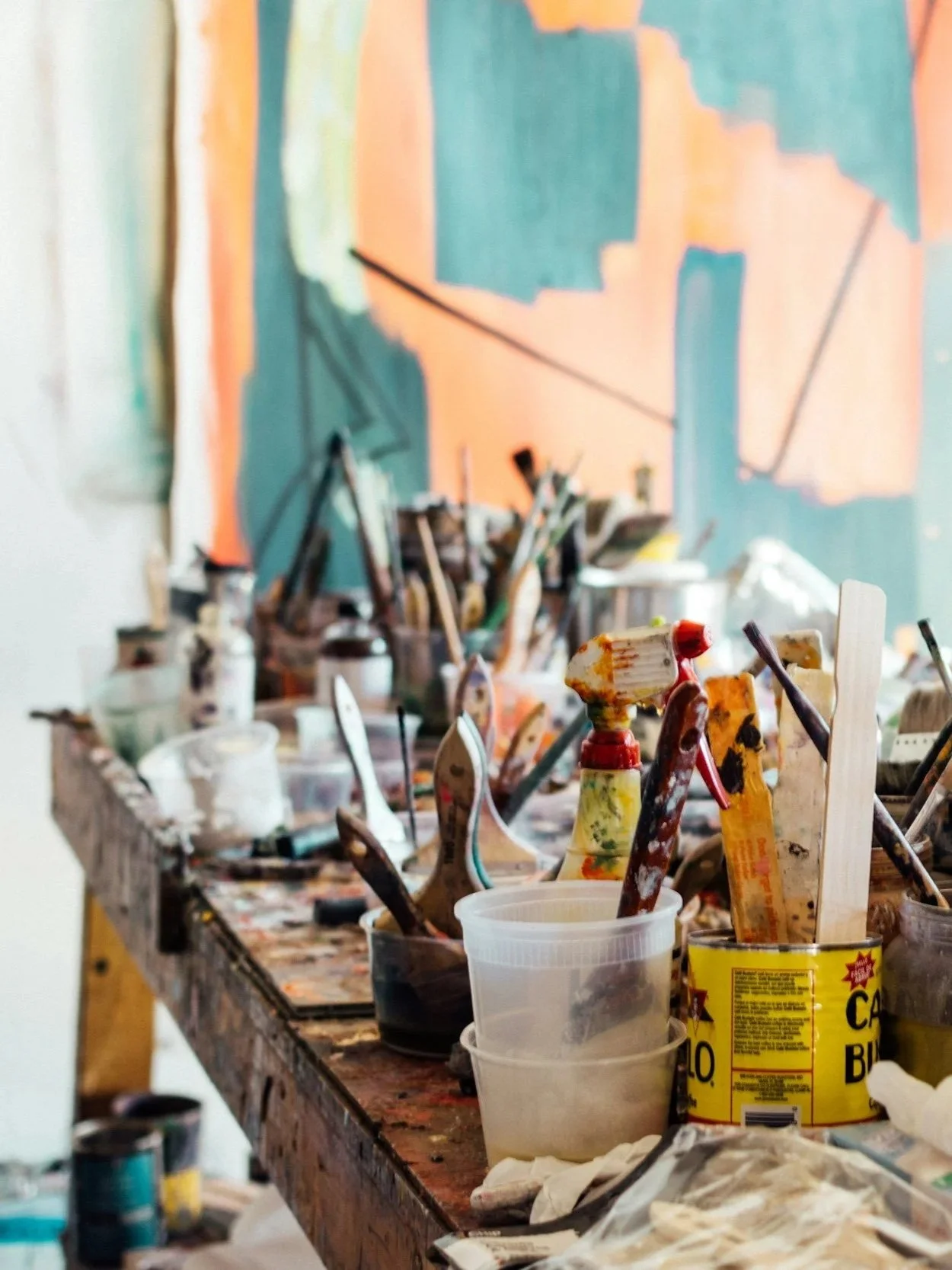 A cluttered artist's workbench with paint bottles, brushes, and painting supplies, with an abstract colorful painting in the background.