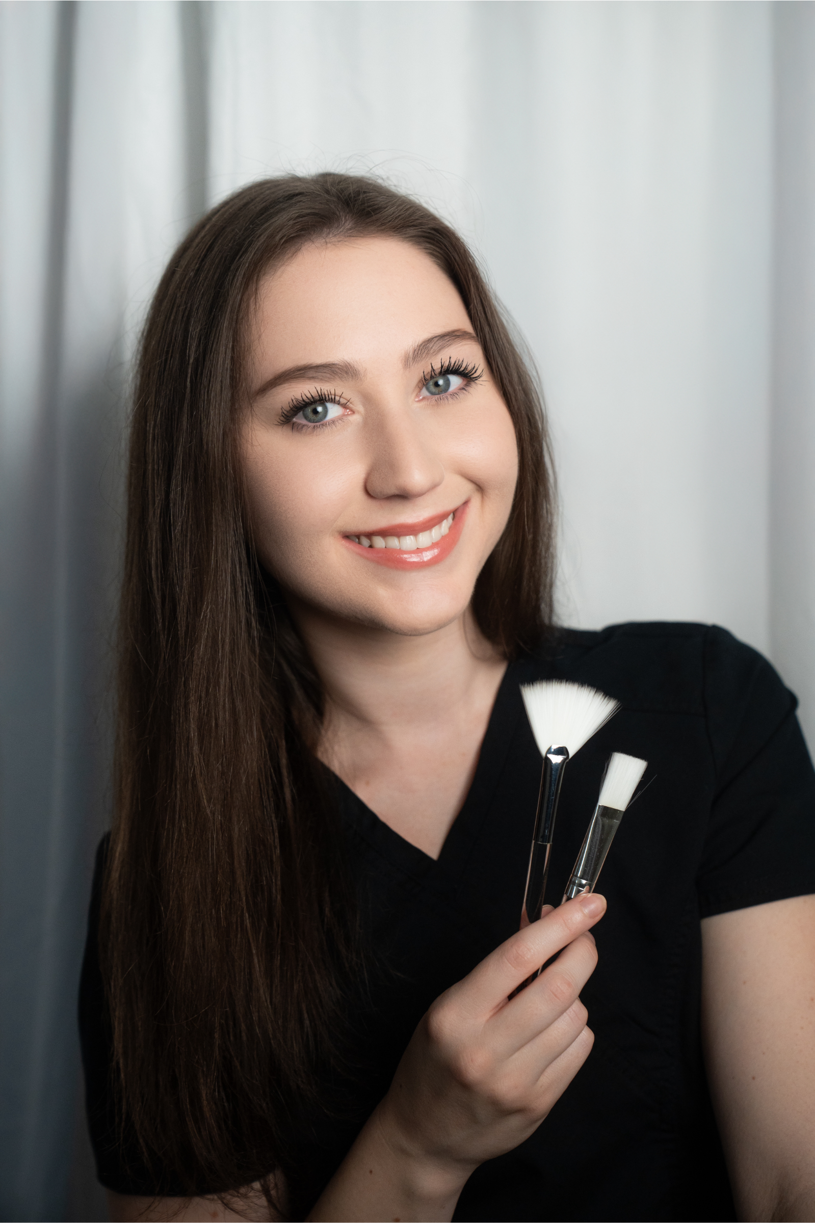 A woman with long brown hair and blue eyes holding three makeup brushes, smiling at the camera, standing against a gray backdrop.