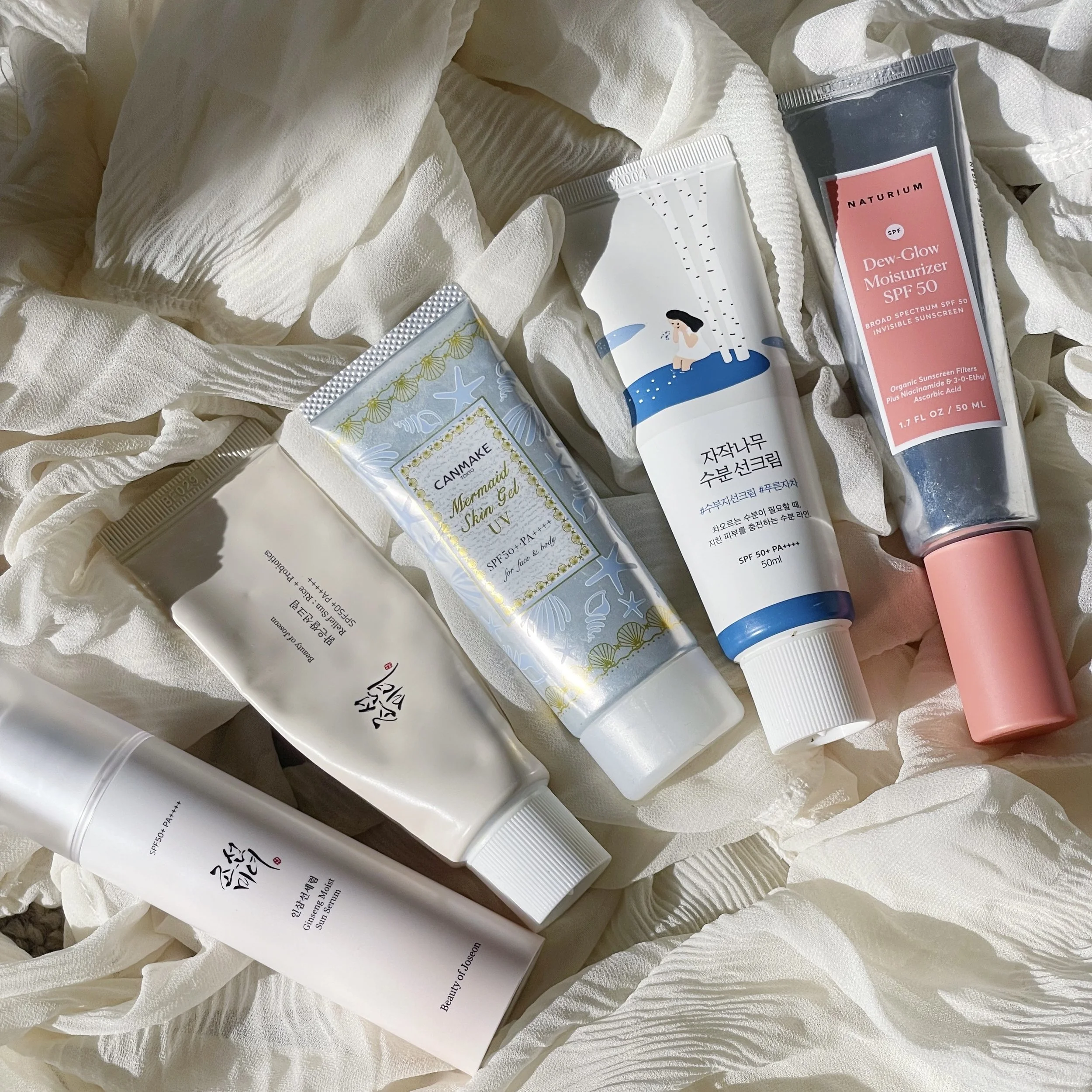 A collection of five skincare products arranged on a white, textured fabric background. The products include moisturizers and sunscreens in various packaging.