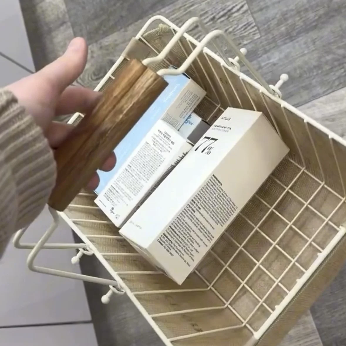 A person holds a wooden-handled brush over an empty white wire basket containing cereal boxes on a wooden surface.