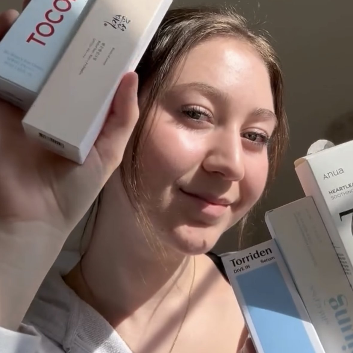 A smiling young woman with brown hair holding skincare product boxes, including TOCO and Anua brands, close to her face.