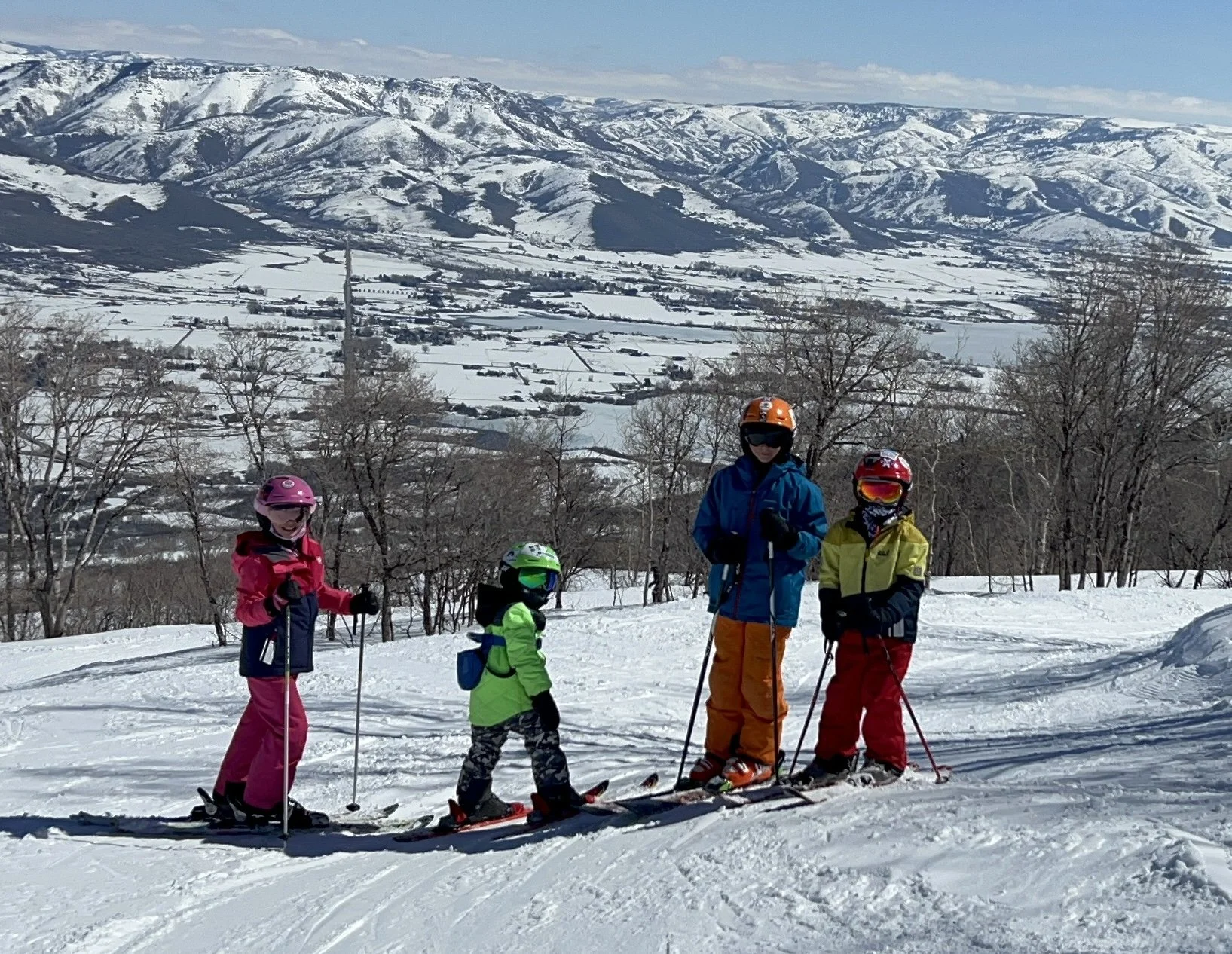 Four children dressed in colorful winter ski gear standing on a snow-covered slope with a scenic view of snow-capped mountains and a valley in the background.