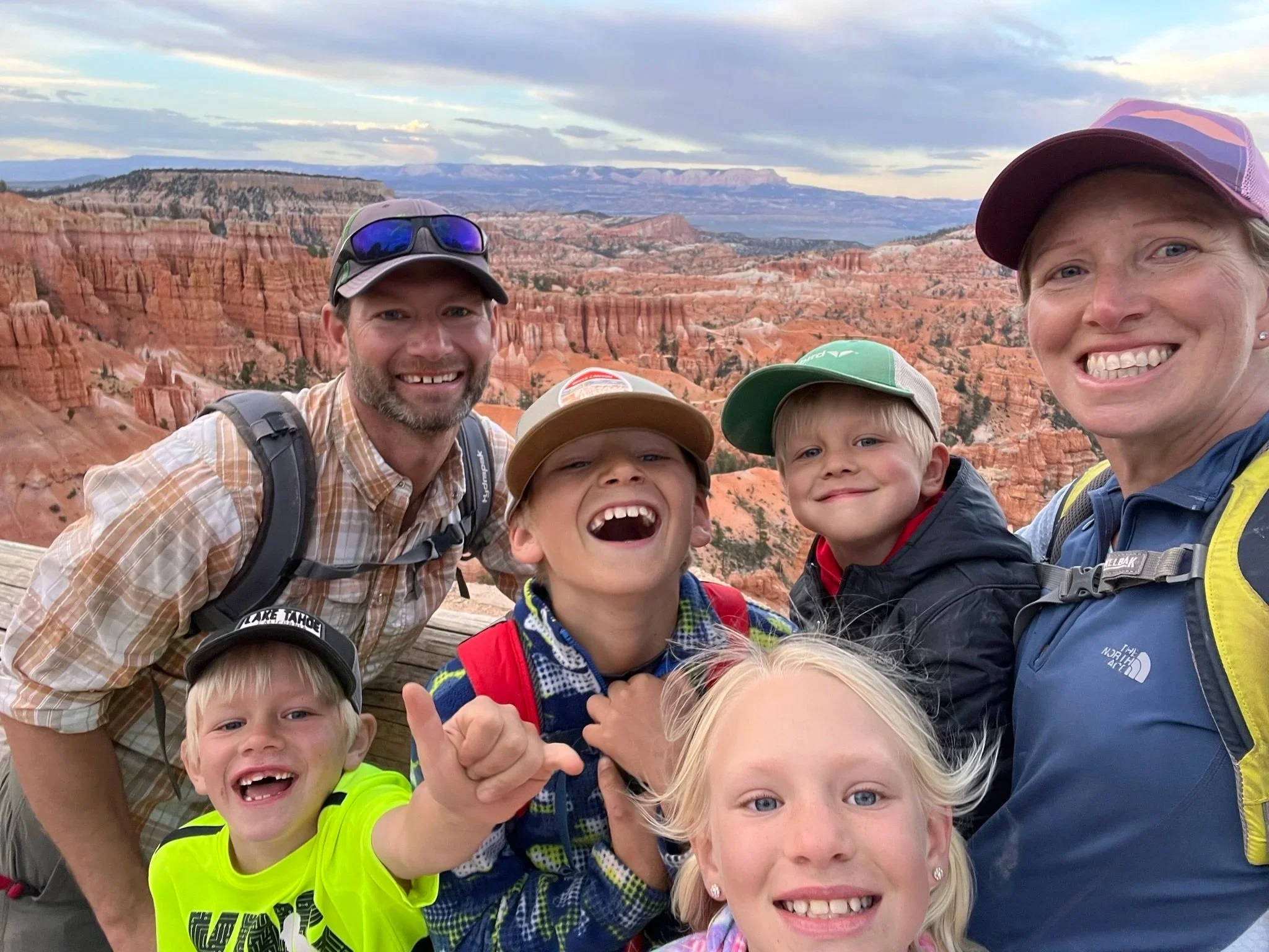 Family of six taking a selfie with a canyon landscape in the background, including red rock formations and distant mesas, during a hike or outdoor adventure.