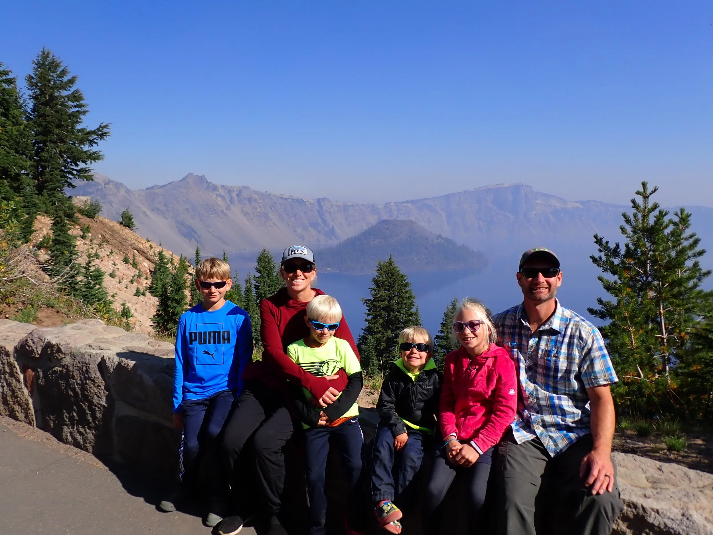 A family of six posing outdoors in front of a mountain and lake scene during daytime. The family consists of two adults and four children, all wearing sunglasses and casual outdoor clothing, sitting on a rock ledge with trees around them.