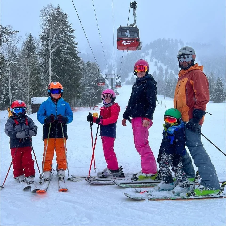 Group of six people, including children and an adult, in winter gear with helmets and ski goggles, standing on skis in a snowy landscape with ski lift and trees in the background.